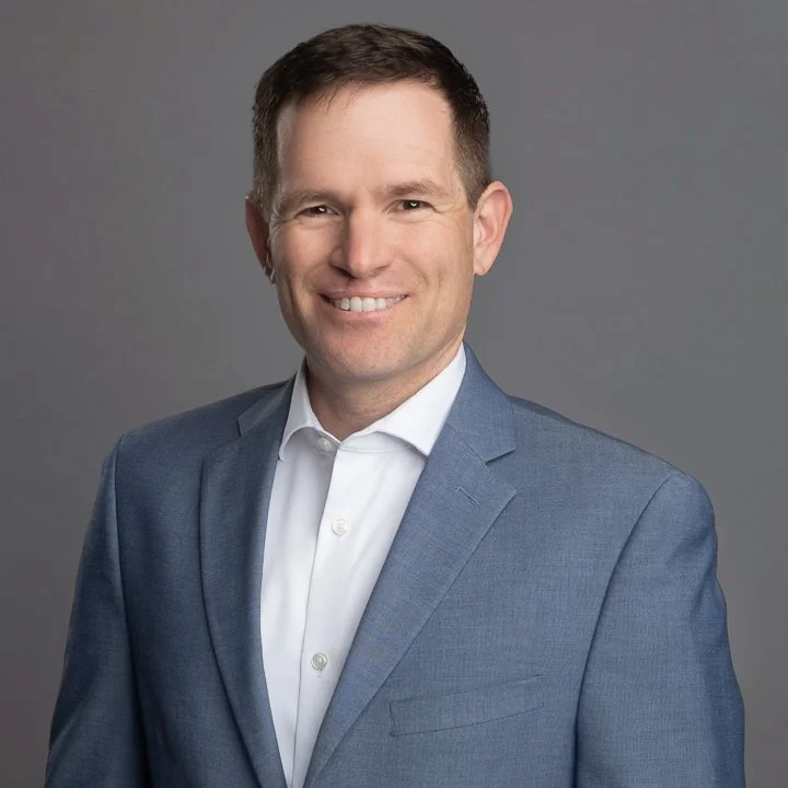 Professional portrait of a smiling man in a blue suit and white shirt against a gray background.