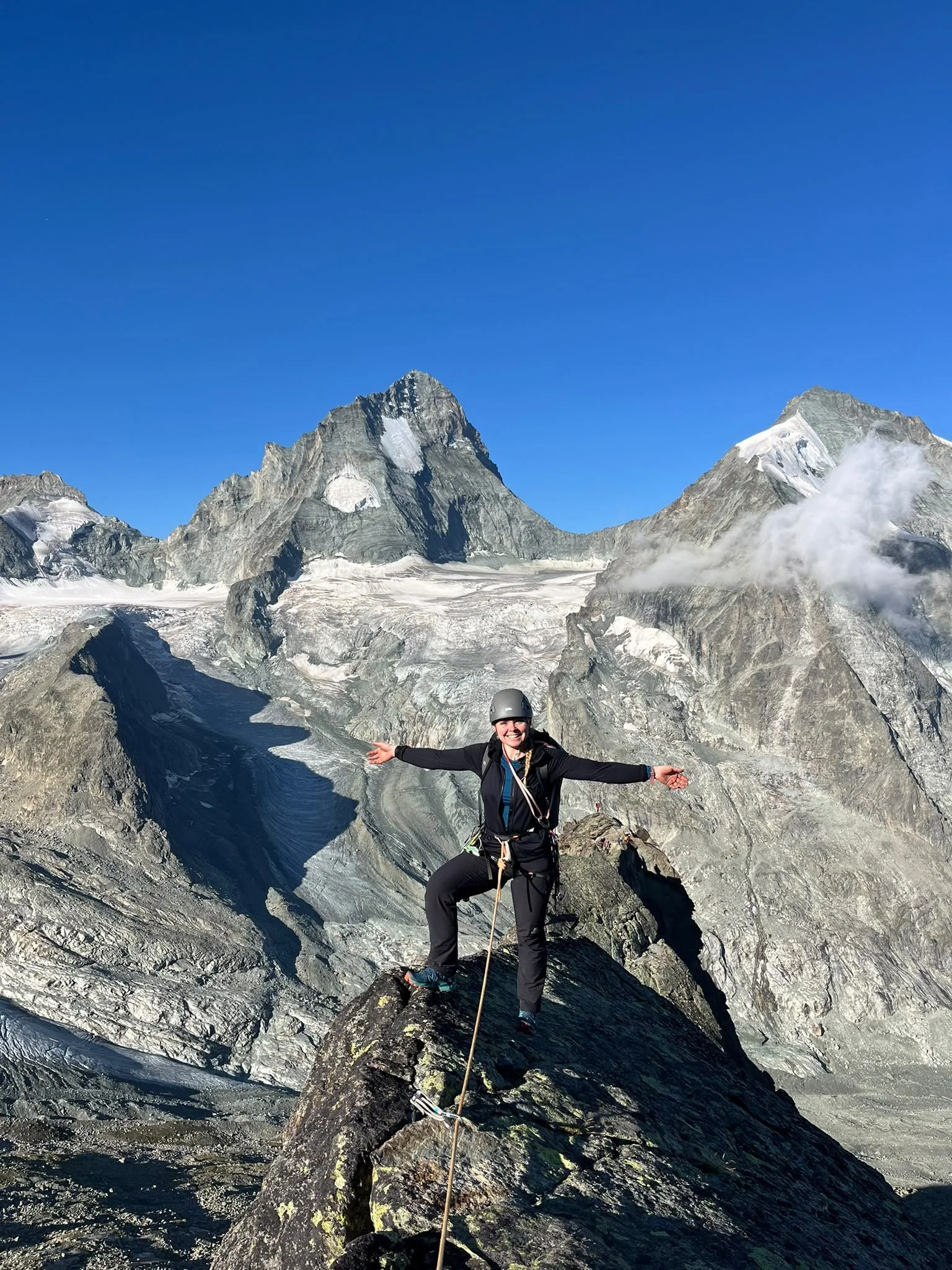 A woman wearing outdoor gear, a helmet, and a harness standing on a narrow rocky outcrop with her arms outstretched, with glacier-covered mountains in the background under a clear blue sky.