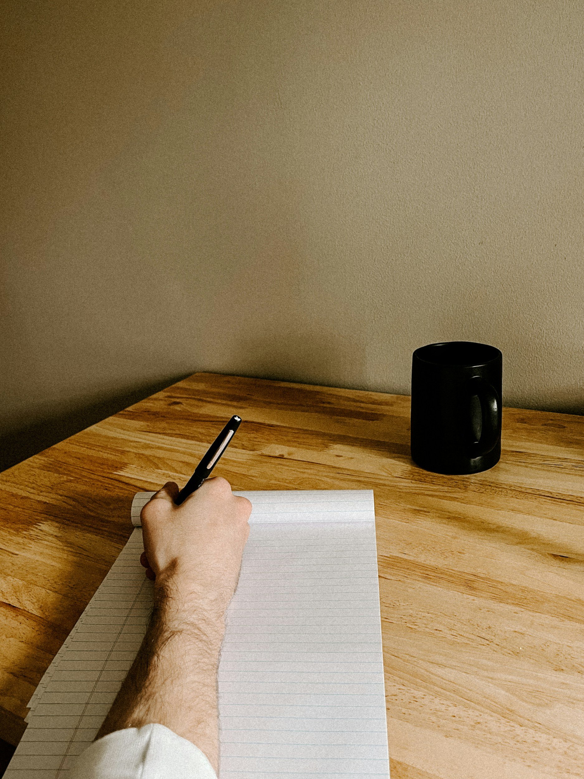 A person writing in a lined notebook with a black pen on a wooden table, a black coffee mug, and a blank wall in the background.