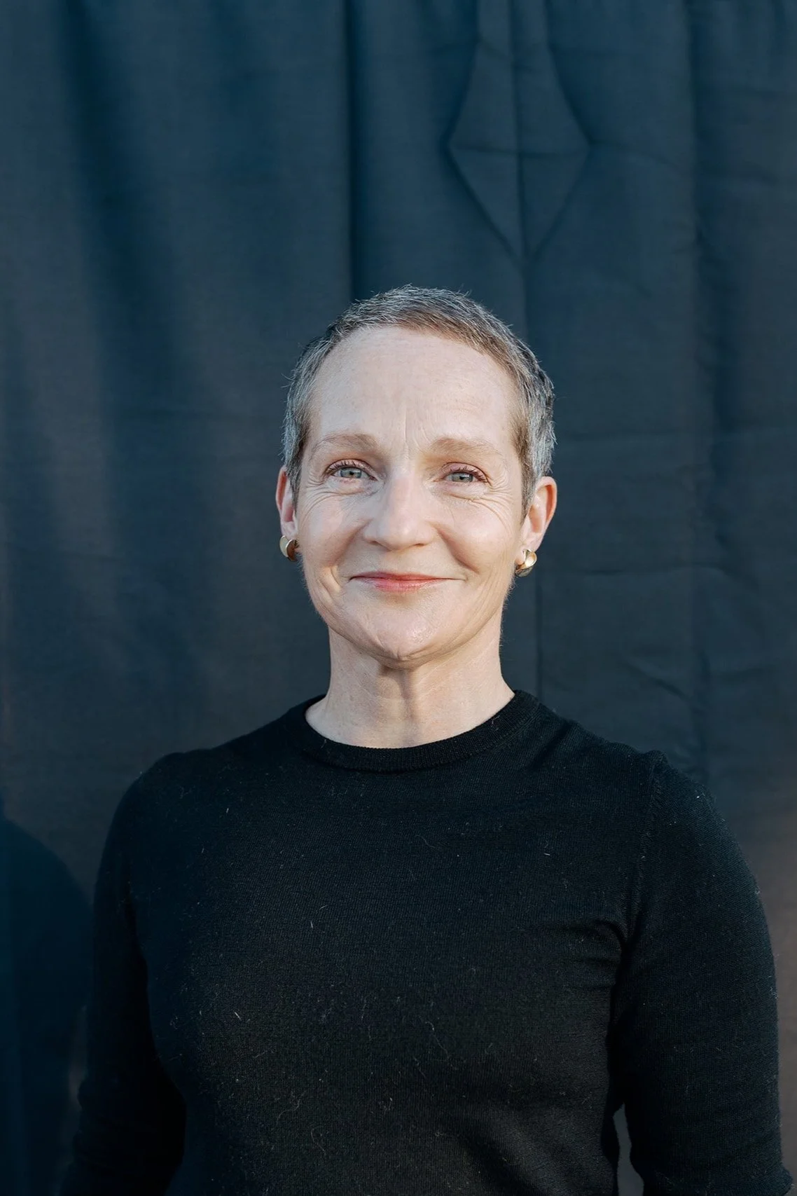 A middle-aged woman with short hair, wearing a black long-sleeve shirt and gold earrings, standing in front of a black background with a geometric design.