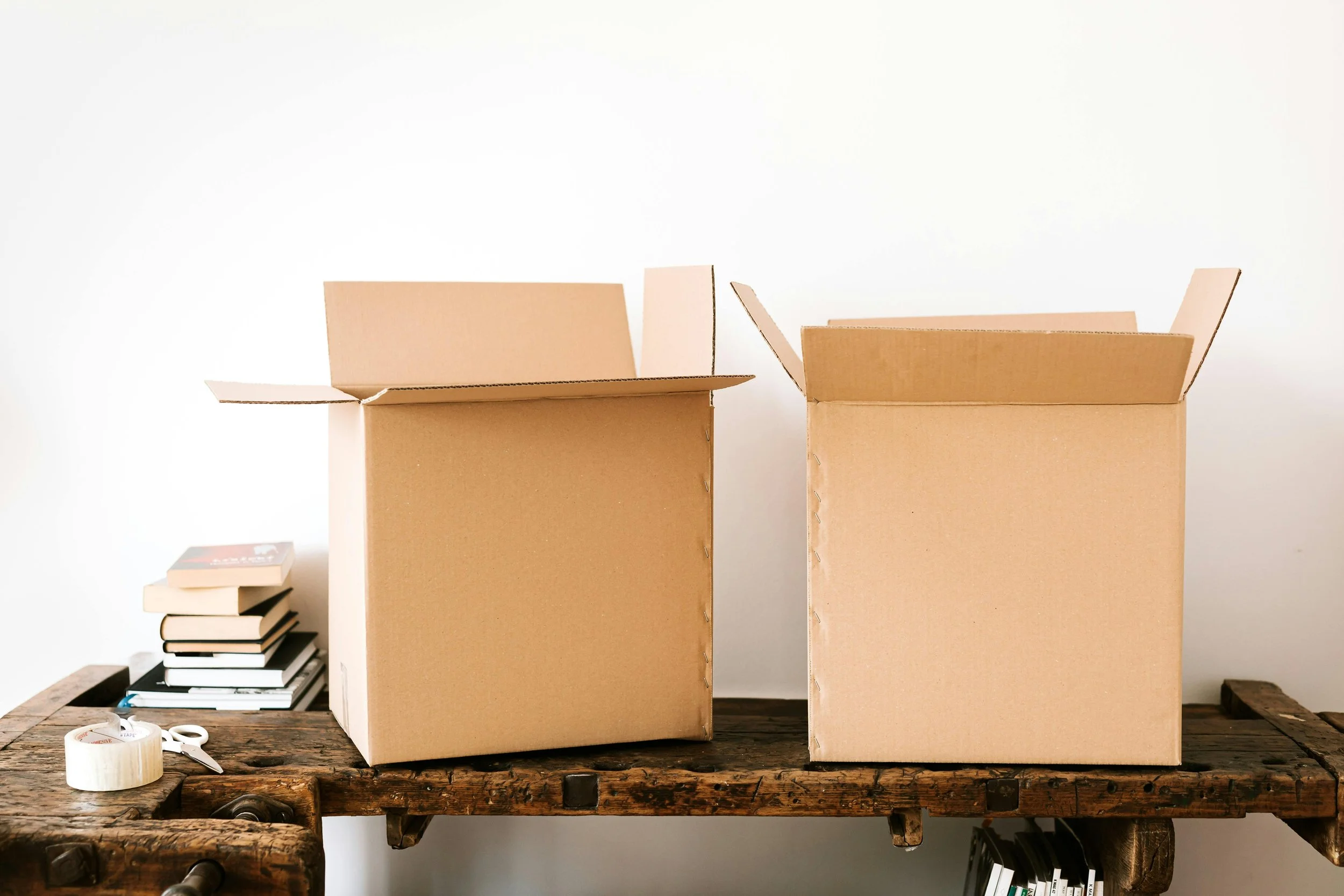 Two cardboard boxes open on a rustic wooden table, with a stack of books and packing tape nearby, against a plain white background.