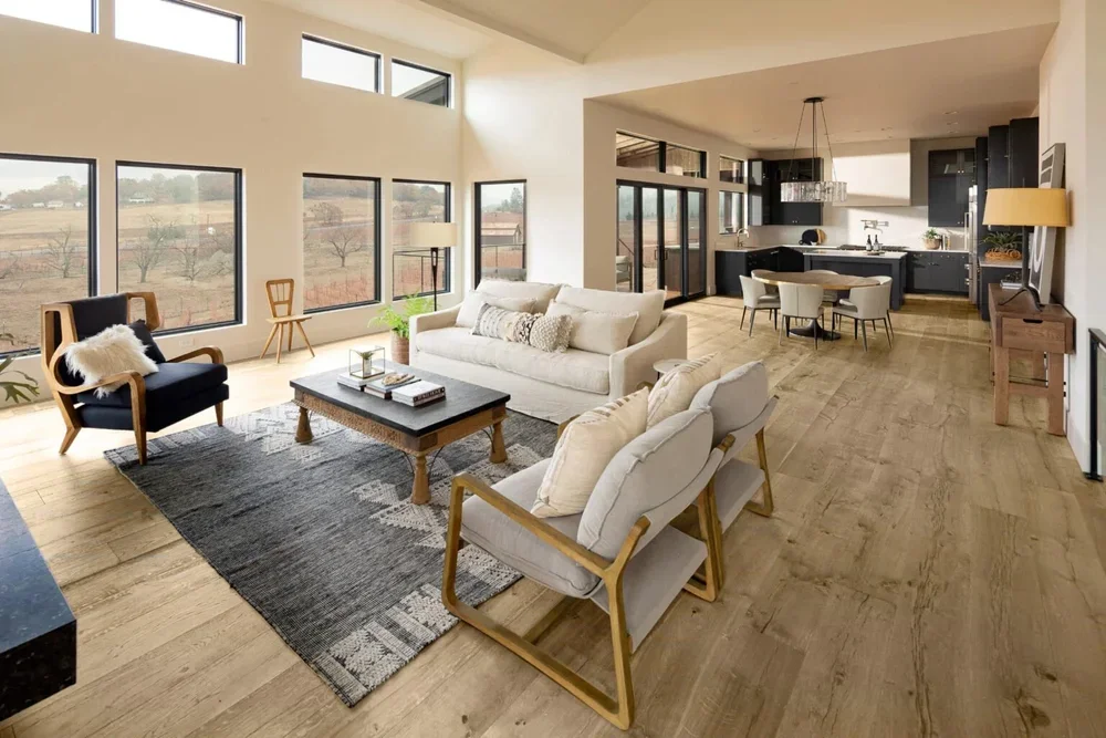 Open-concept living room and kitchen with large windows, modern furniture, and wood flooring.