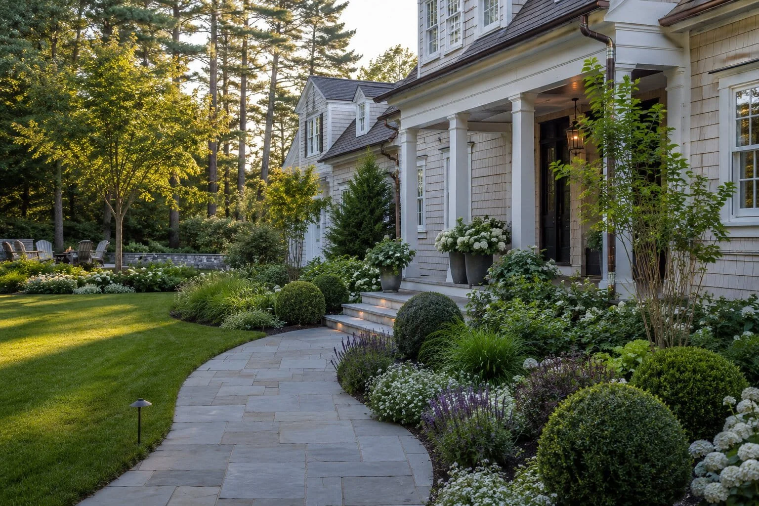 A landscaped front yard with a curved stone pathway, lush green grass, and well-maintained shrubs and flowers. The house has white siding, columns at the entrance, and potted plants, with trees and more greenery in the background.