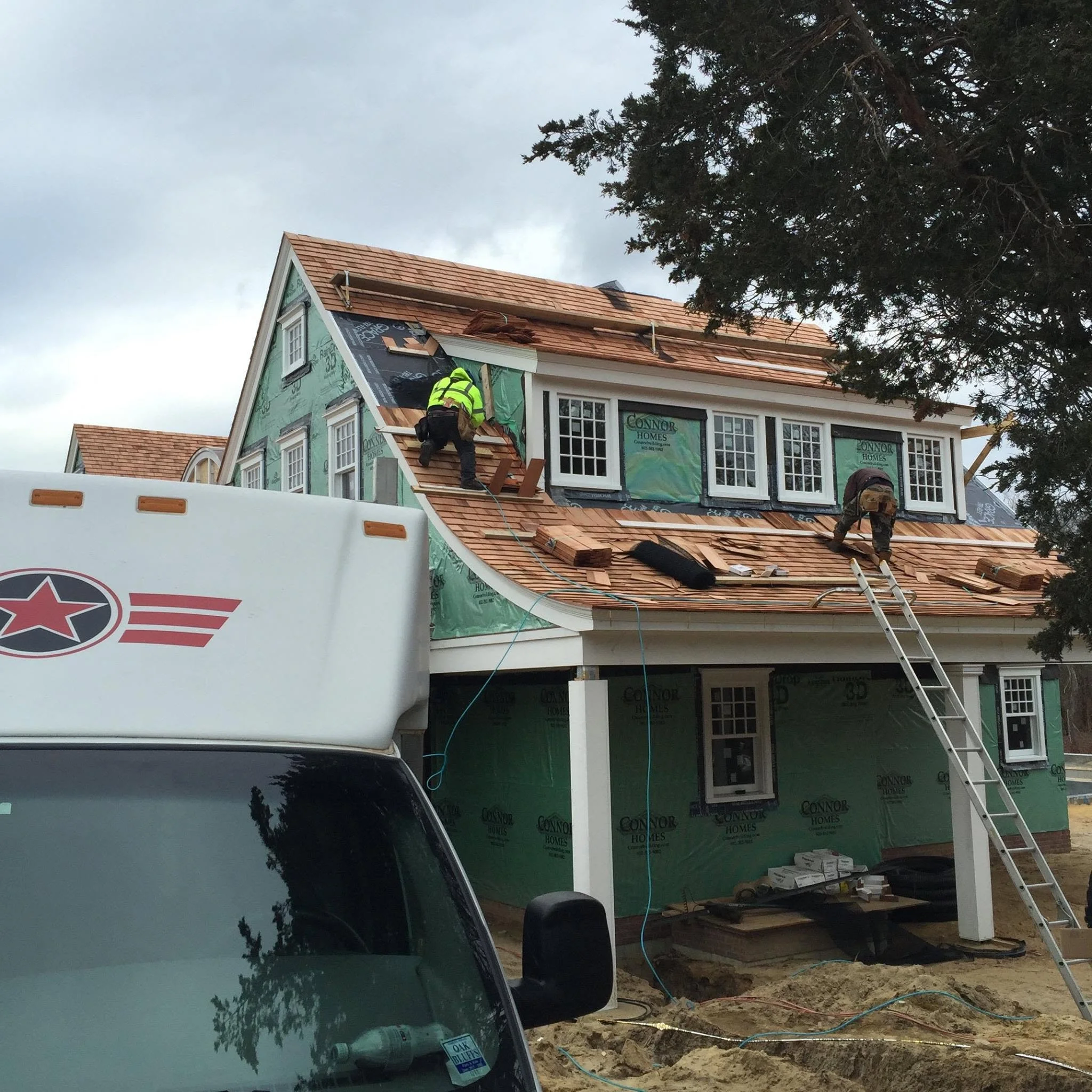 Construction workers building or renovating a house, working on the roof with shingles, ladders, and construction materials around.