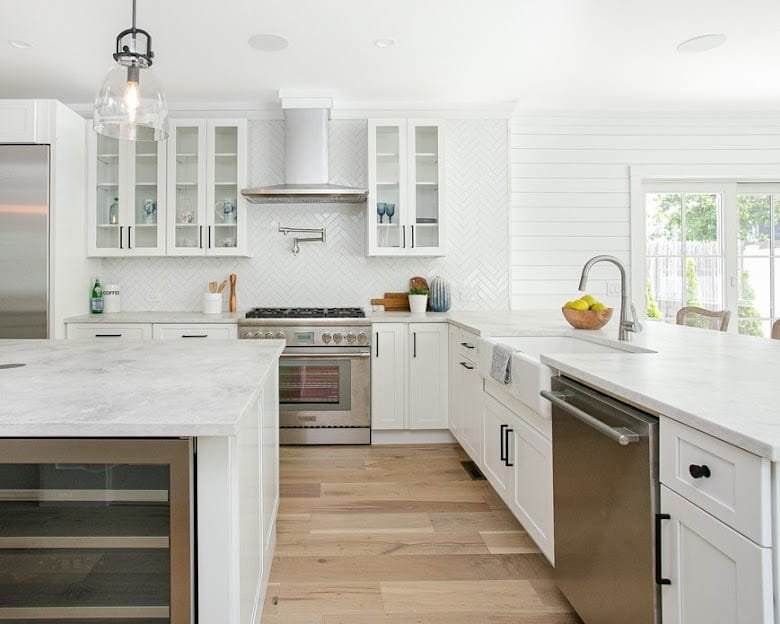 Modern white kitchen with marble countertops, stainless steel appliances, glass-front cabinets, and a large island. There is a window with natural light, a bowl of fruit, and wooden flooring.