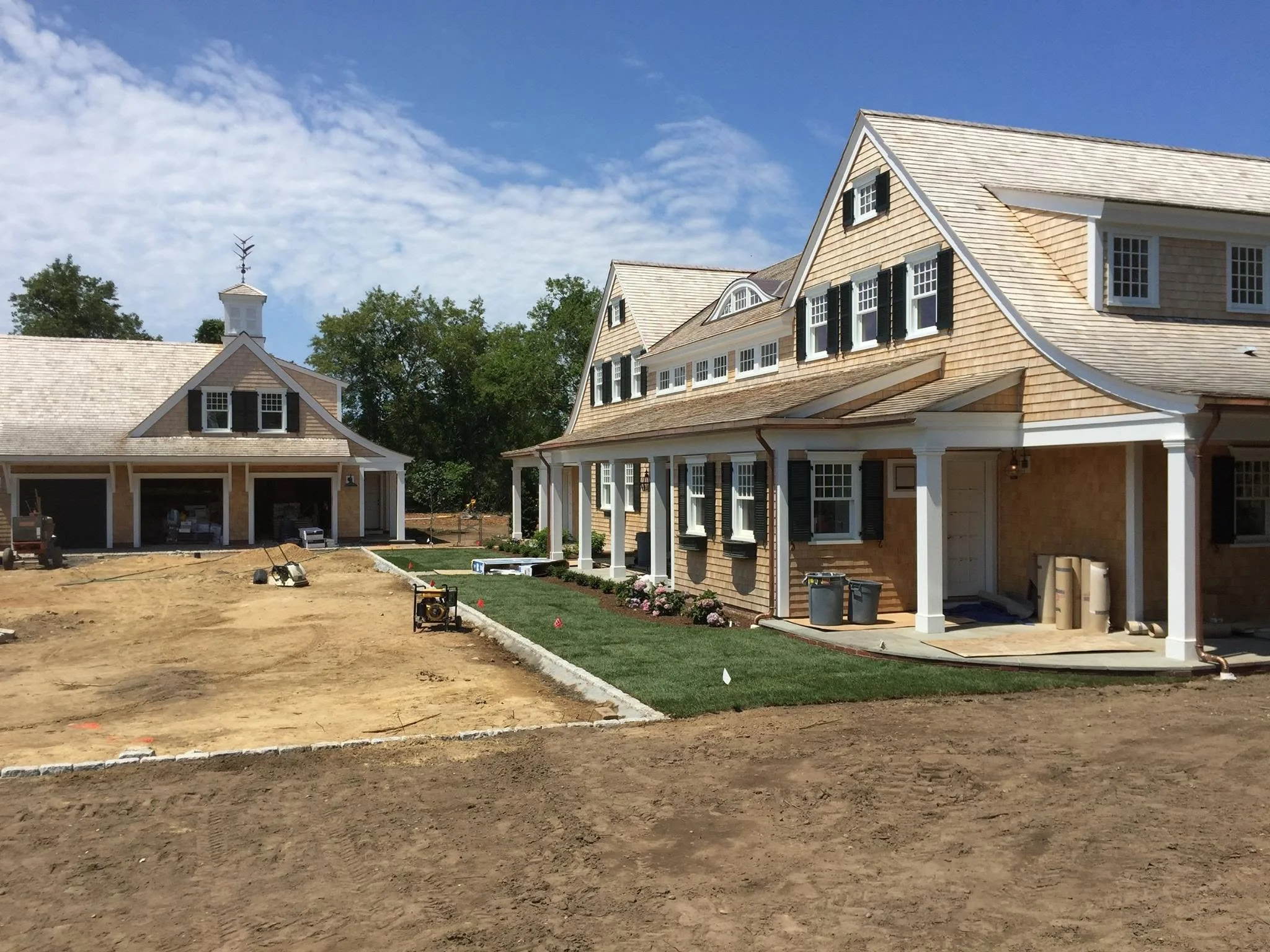 Under construction of a house, front yard with a strip of green grass and a concrete border, partially built house with beige siding, black shutters, and white trim, a small garage with open doors and construction tools in the yard, trees in the background, blue sky with some clouds.
