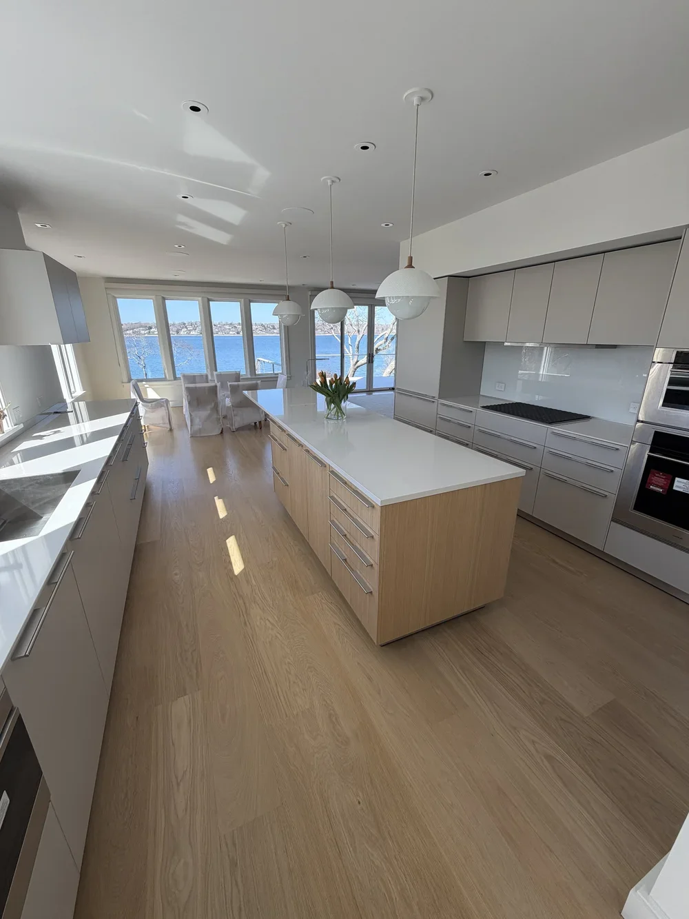 Modern kitchen with white island, light wood cabinetry, and a view of a body of water through large windows.