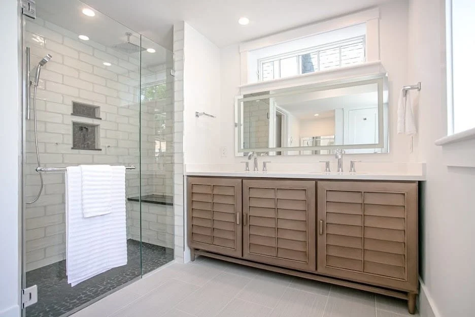 Modern bathroom with a glass-enclosed shower area featuring white subway tiles, a built-in niche, and a rainfall showerhead. A wooden vanity with shutter-style cabinet doors, a large mirror, and a double sink countertop. There are windows with white trim above the vanity and natural light coming into the room.