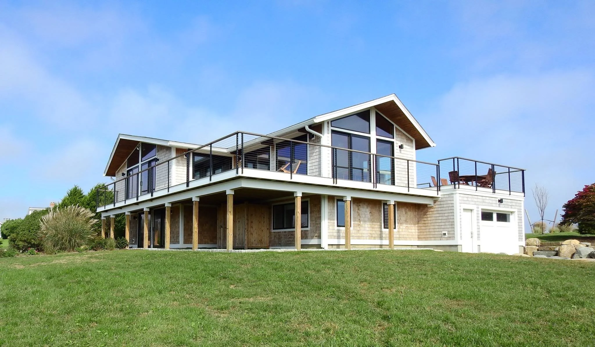 Modern two-story house with a large balcony, white and gray exterior, and glass doors, set against a blue sky with some trees and greenery in the yard.