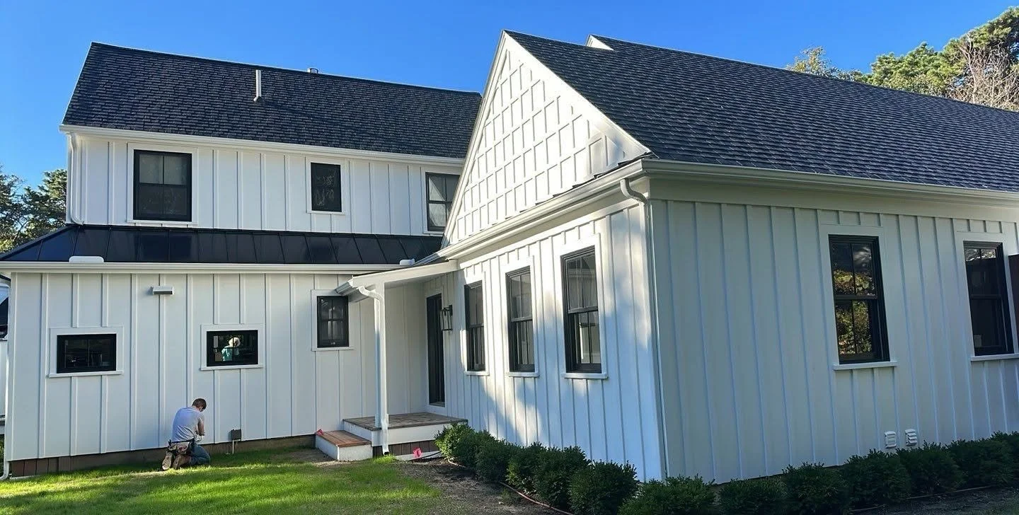 A white two-story house with black window frames and a black roof, featuring vertical siding and a small porch with steps.