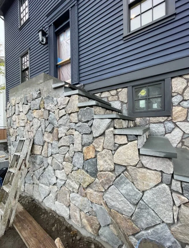 Photo of a house with a stone foundation and blue siding, showing a small set of concrete steps leading to a door, with a window nearby.