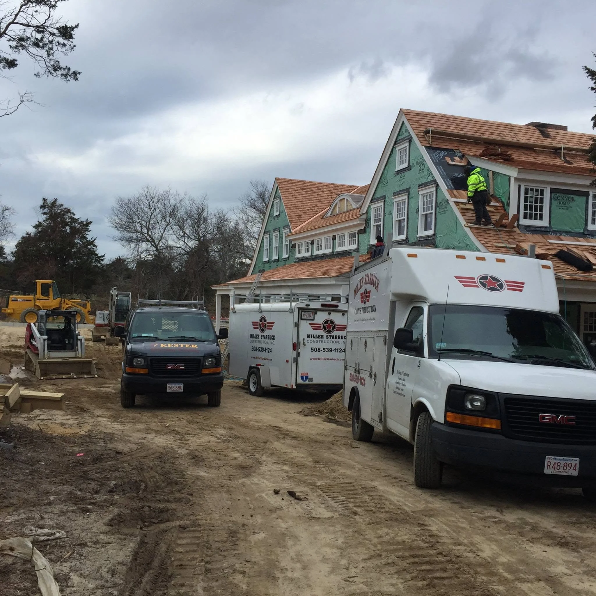 Construction site with a partially built house, construction workers, and equipment under a cloudy sky.