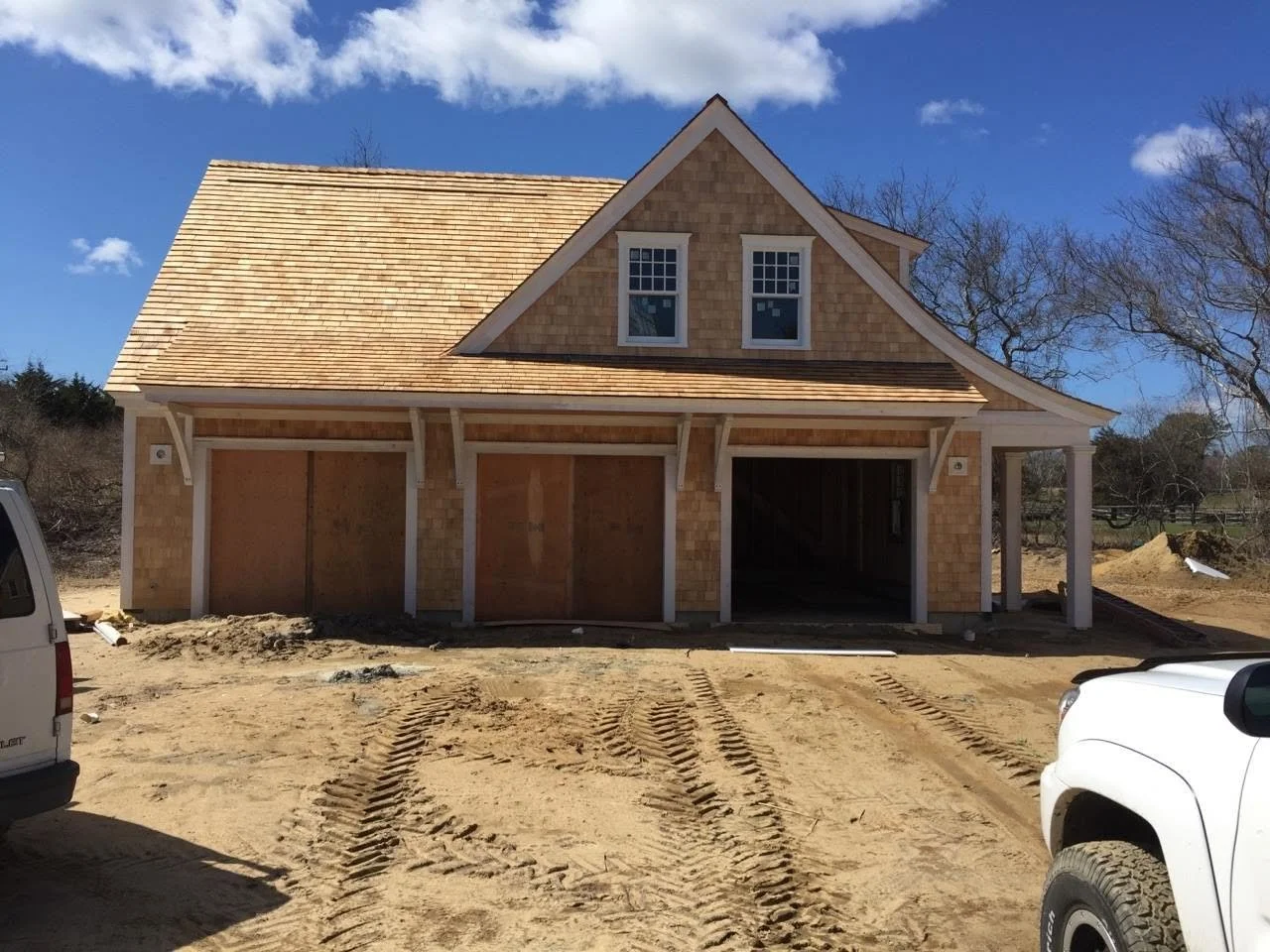 Under construction two-story house with a wooden roof on a dirt lot, surrounded by vehicles and trees in the background.