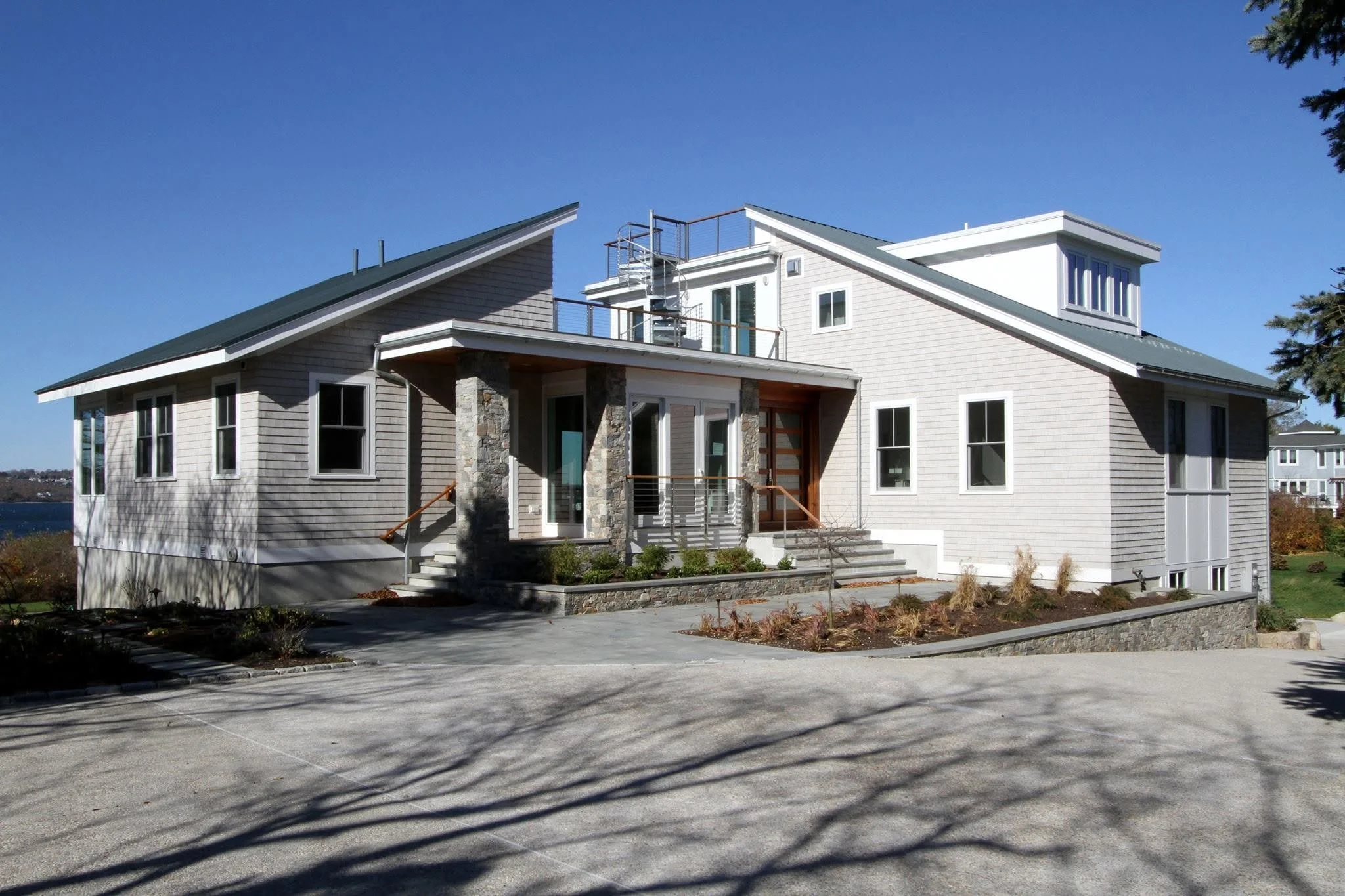 A modern two-story house with beige siding, stone accents, and a metal roof, with an outdoor spiral staircase leading to the roof deck. The house features multiple windows and a small front porch with stairs, surrounded by a parking lot and some landscaping, under a clear blue sky.
