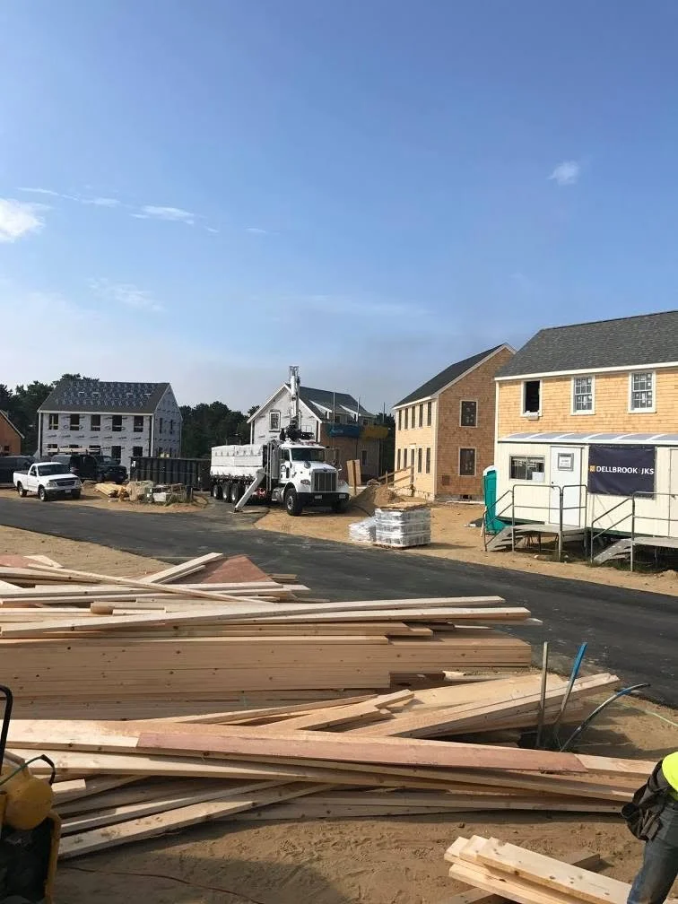 Construction site with stacks of wooden planks, a white dump truck, and partially built houses under a clear blue sky.