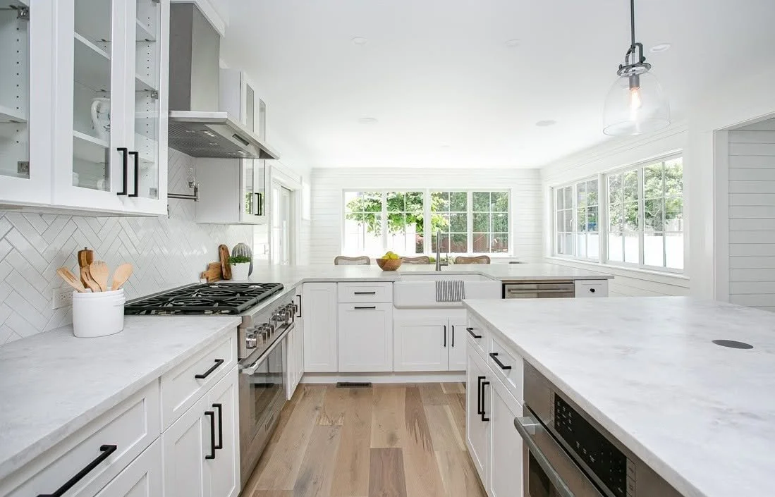 Bright white kitchen with large windows, white cabinets, marble countertops, stainless steel appliances, and light wood flooring.