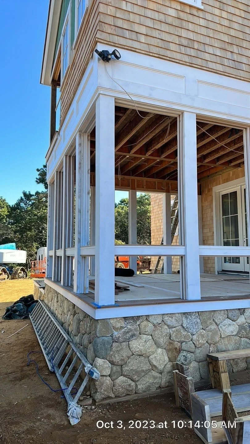 Construction site of a house with stone foundation and wooden framing, partial exterior wall with siding, ladder, and construction tools visible.