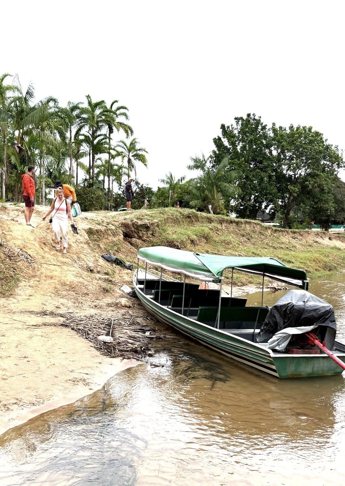 People walking along a dirt riverbank with a green boat anchored nearby, lush trees in the background, overcast sky in Mutum Village.