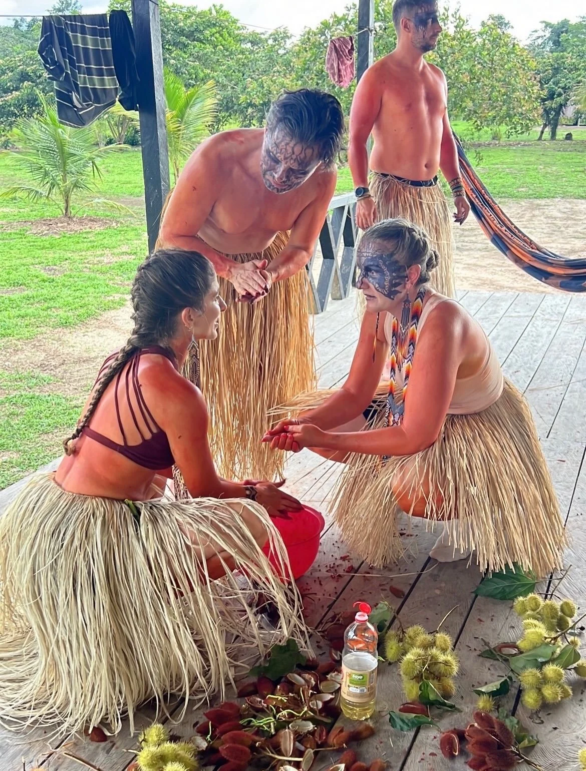 Yawanawa people participating in a traditional ceremony outdoors, with some wearing grass skirts, face paint, and holding ritual items, surrounded by floral and natural elements.