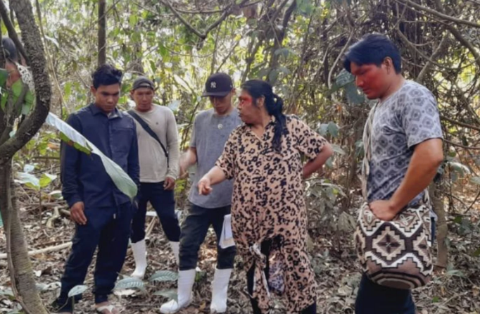 A Yawanawa woman in leopard print clothing showing a plant or leaf to four young men in a dense forest or jungle.