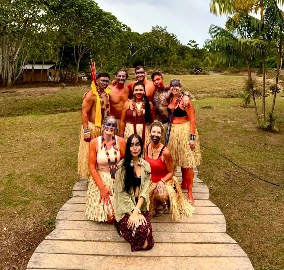 Group of people dressed in traditional Polynesian or Pacific Islander attire, standing on a wooden pathway outdoors with trees and grassy area in the background.