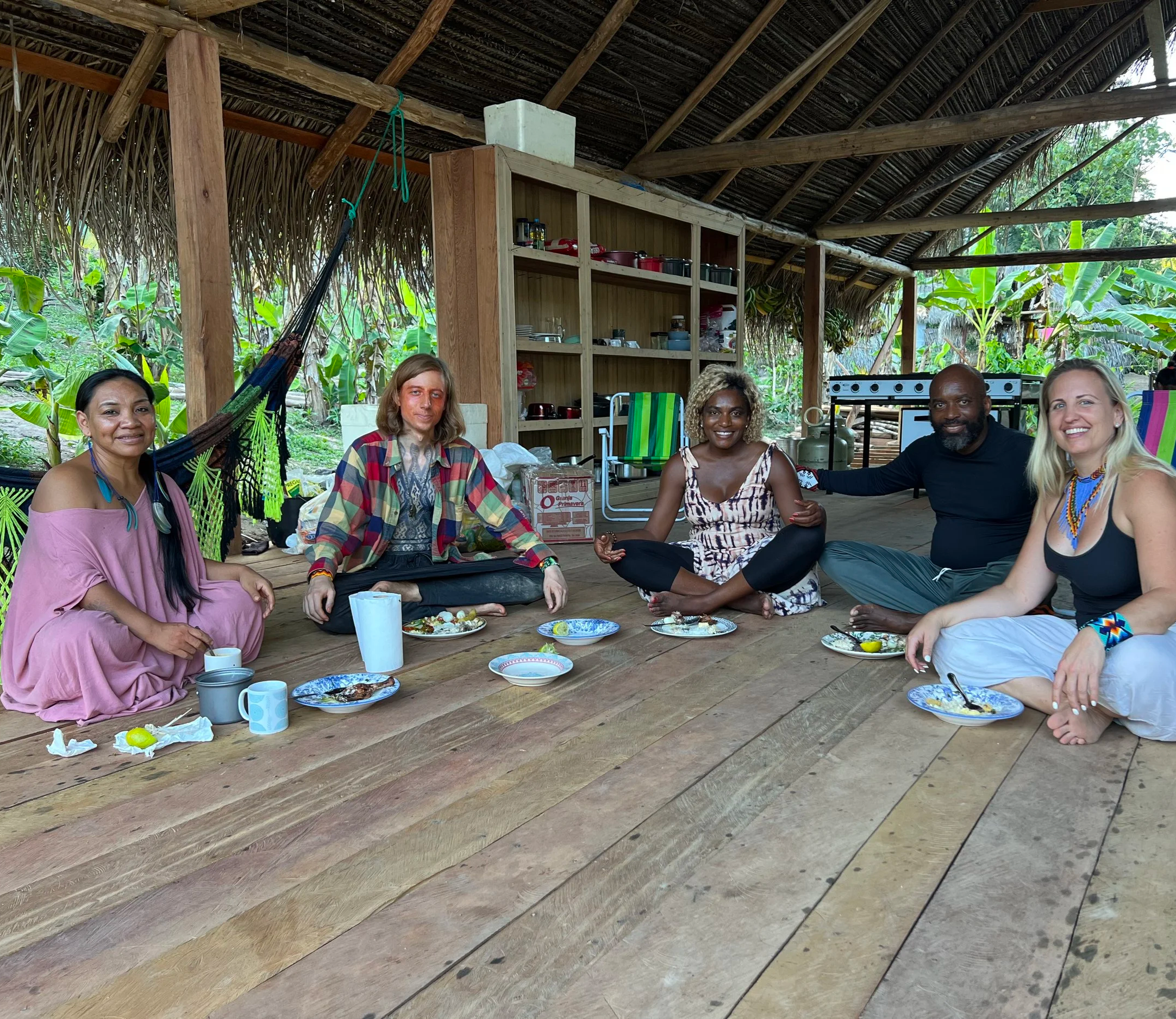 Group of five people sitting on a wooden floor, with plates of food and drinks, in an outdoor, open-air shelter with a thatched roof surrounded by greenery in Mutum Village.