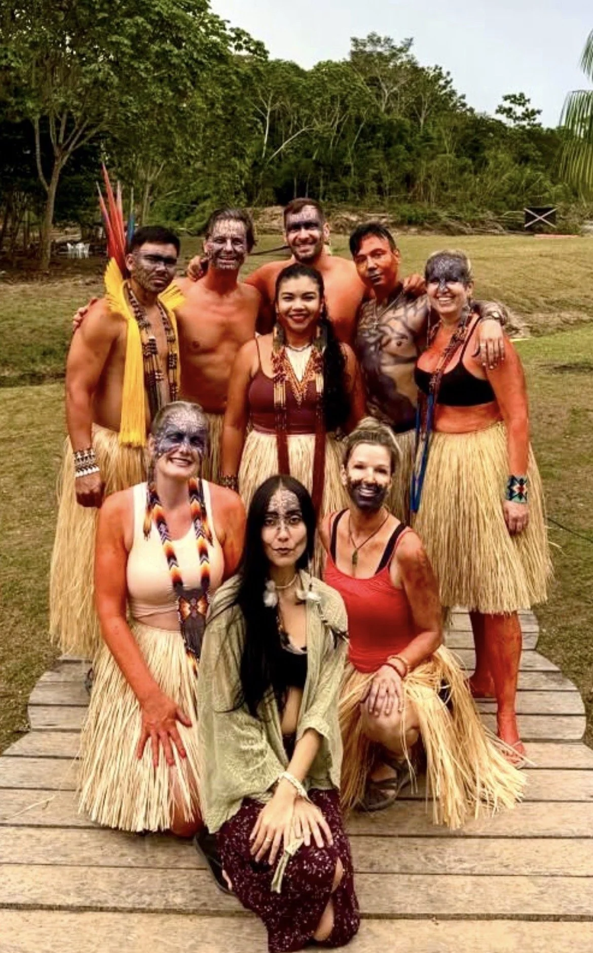 Group of people dressed in traditional Yawanawa attire, including grass skirts and face paint, standing outdoors on a wooden platform with a grassy field and trees in the background.