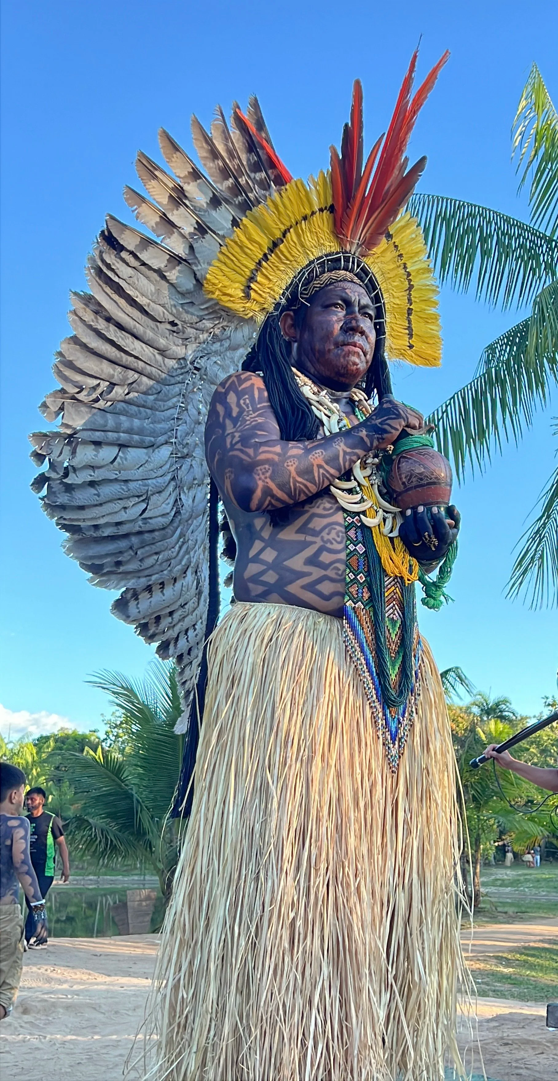 Indigenous man dressed in traditional attire with a large feathered headdress, body tattoos, and holding a clay pot, standing outdoors with trees and a clear blue sky in the background.