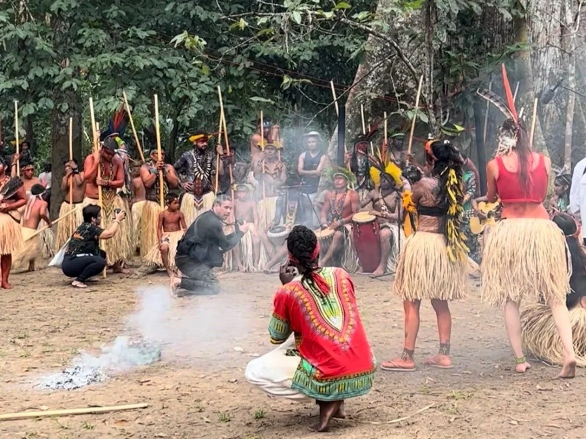 Indigenous people dancing and playing traditional drums in a forest, wearing grass skirts, feathers, and colorful clothing.