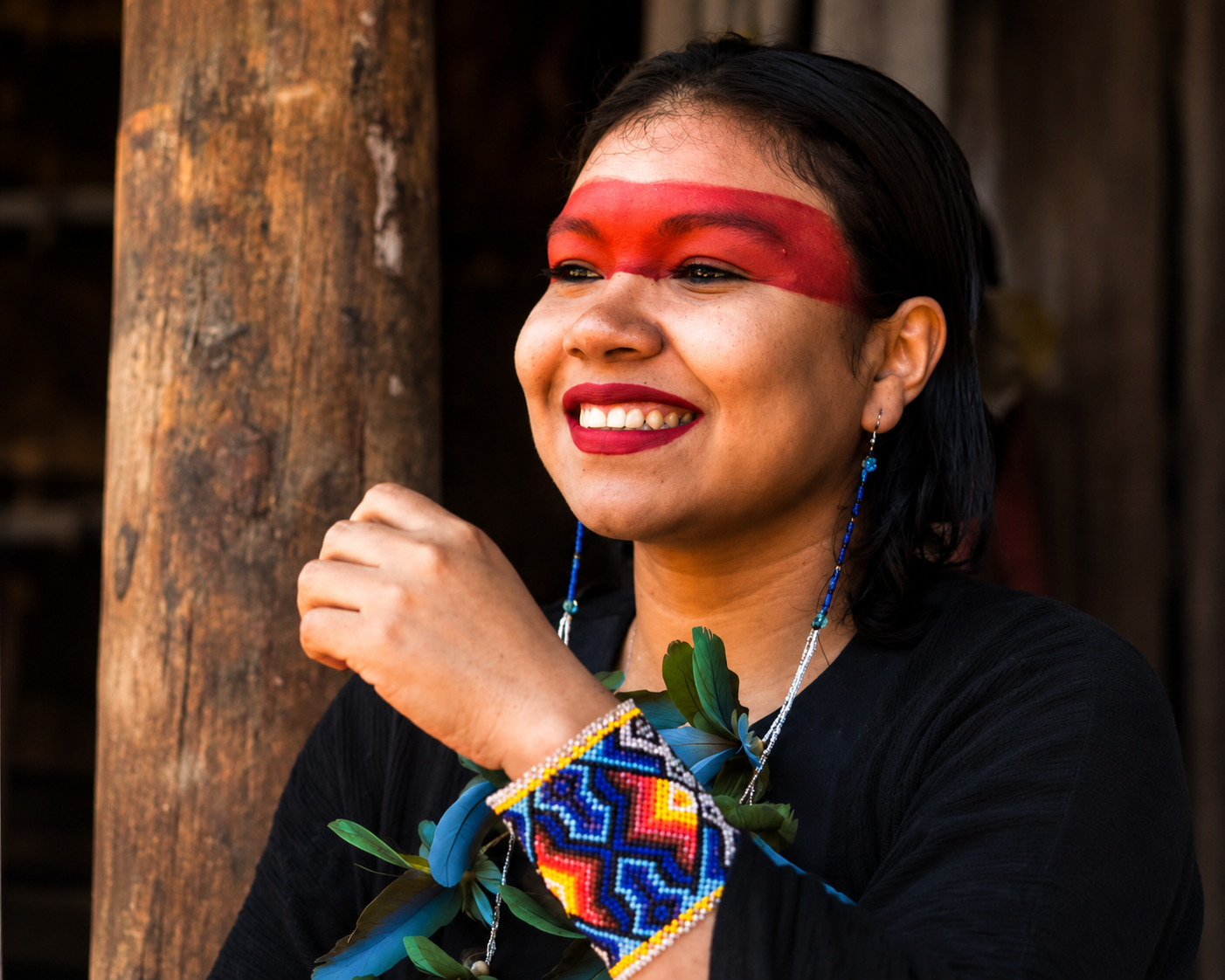 A Yawanawa woman with dark hair decorated with traditional jewelry and face paint, standing near a wooden post.