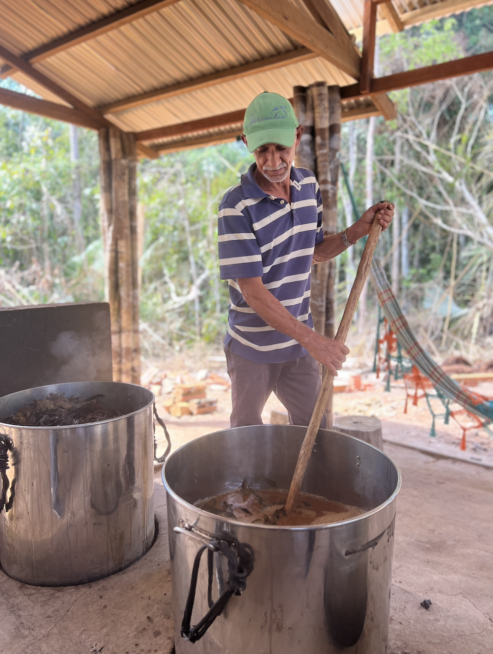 An Yawanawa man in a green cap and striped polo shirt stirs a large pot of food in a rustic outdoor kitchen with wooden beams and a hammock in the background.