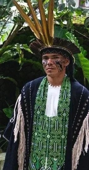 Yawanawá man dressed in traditional indigenous clothing, including a headdress with long feathers, face paint, and a woven garment, standing amidst lush green foliage in Mutum Village.