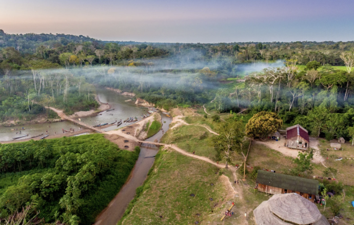 Aerial view of Mutum Village, a lush green landscape with a river, small boats, a rustic house, and a thatched-roof structure, surrounded by trees and fog at dawn or dusk.