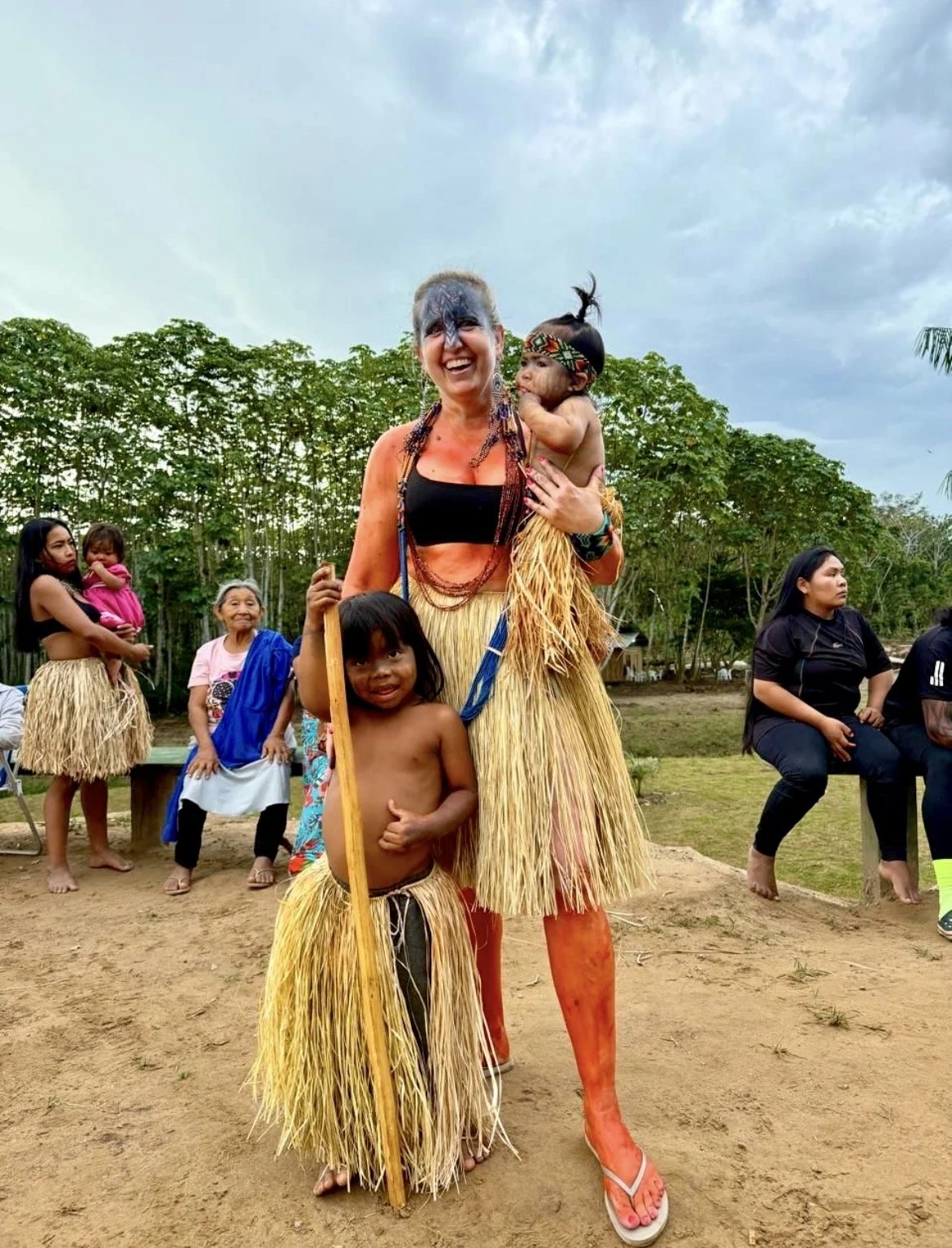 A Yawanawa woman dressed in traditional indigenous attire holding a child, with other children and adults in similar attire and clothing seated outdoors on benches, against a background of trees and a cloudy sky.