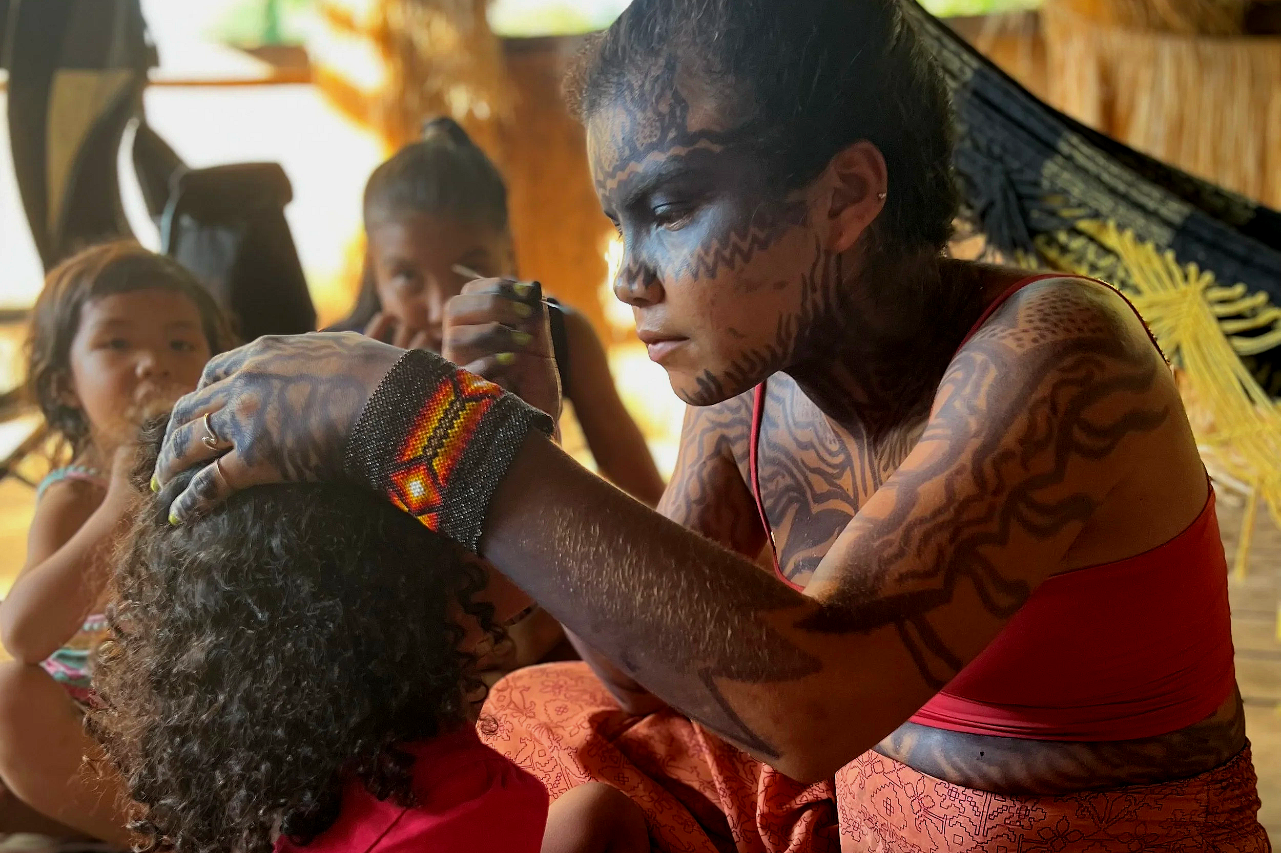 A woman with tribal face paint and tattoos demonstrates a traditional tattooing or body art technique on a young girl with curly hair and beaded jewelry, while others watch in the background, inside a warmly lit wooden structure.