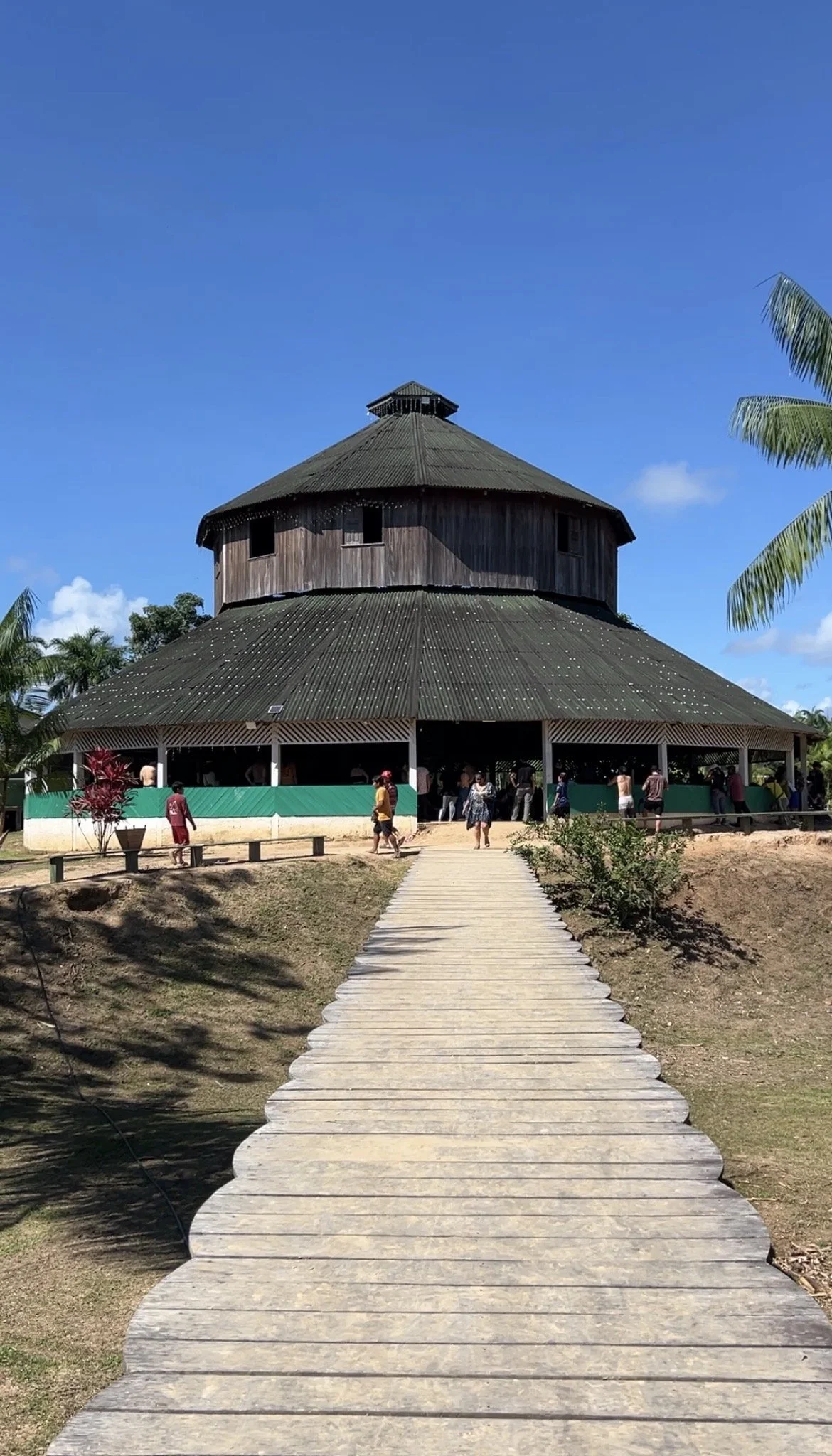 A round wooden building with a conical roof, people walking toward the entrance, wooden walkway, surrounded by tropical plants and palm trees, under a blue sky in Mutum Village.