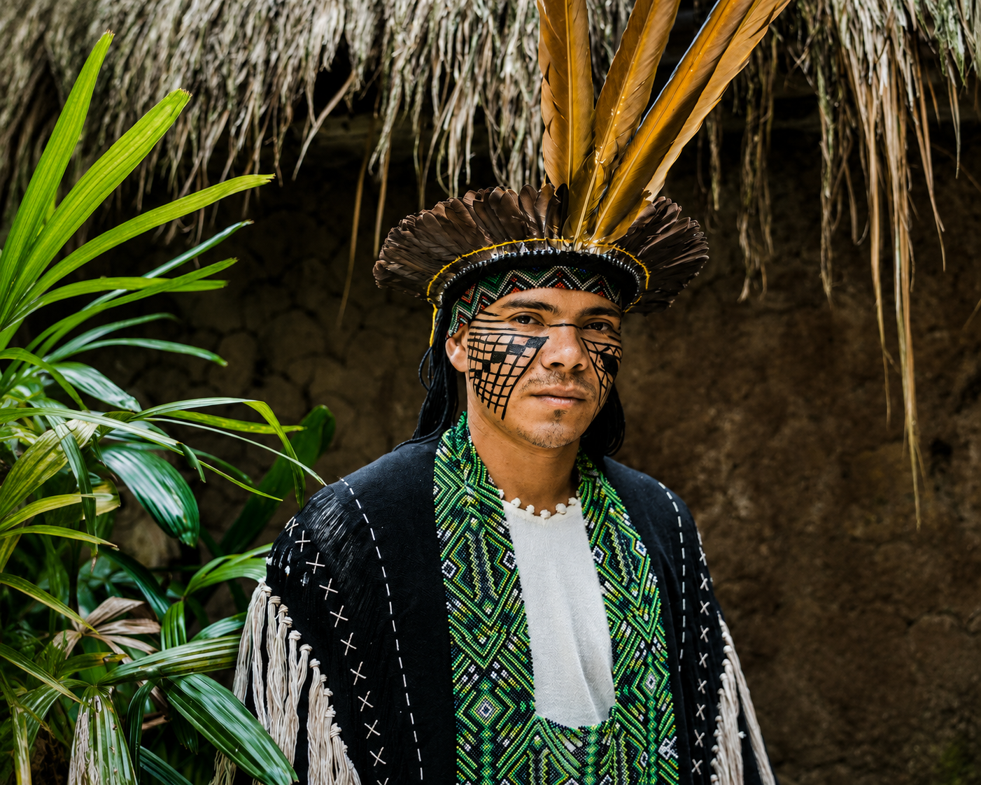 A Yawanawá man dressed in traditional indigenous attire, including a feathered headdress and face paint, standing outdoors surrounded by green plants.