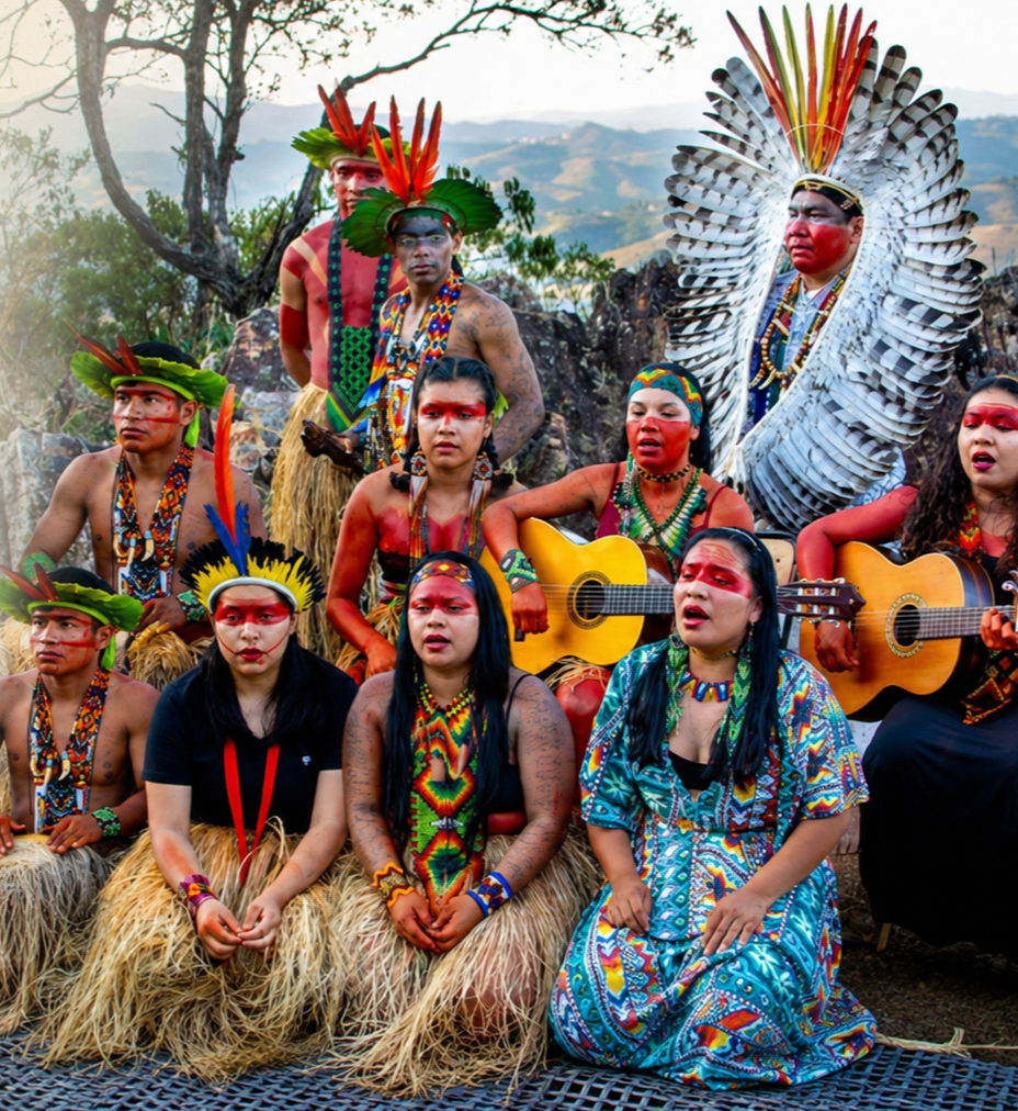 A group of indigenous people dressed in traditional attire, with some wearing feathered headdresses, face paint, and grass skirts. Some members are holding guitars and appear to be performing music outdoors with a scenic landscape in the background.