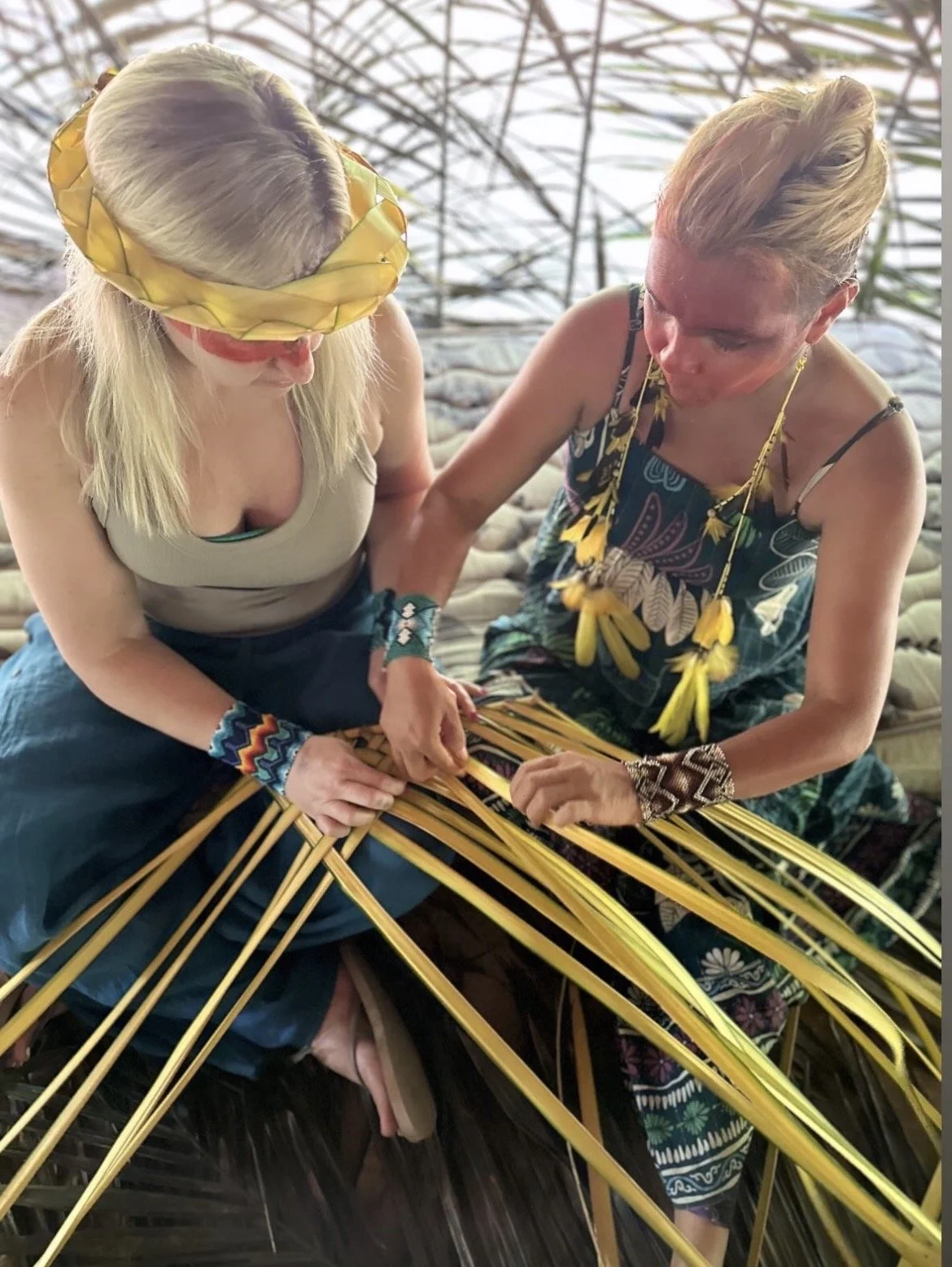 Two Yawanawa women with tribal-inspired accessories and clothing weaving together using yellow strips of material in a greenhouse-like setting.