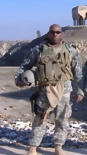 A soldier in camouflage uniform and tactical gear standing outdoors, holding a helmet, with a desert-like landscape and a structure in the background.