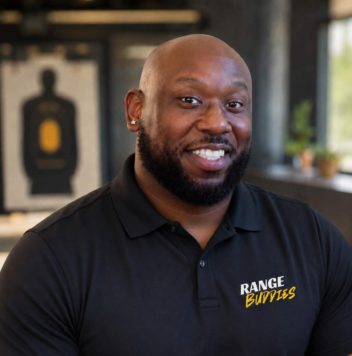 A smiling Black man with a beard, wearing earrings and a black polo shirt with "Range Bubbles" logo, sitting indoors with target practice target and plants in the background.