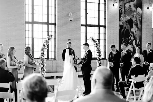 Bride and groom holding hands at the altar during a wedding ceremony in a church, with bridesmaids and groomsmen standing beside them, and guests seated facing the couple.