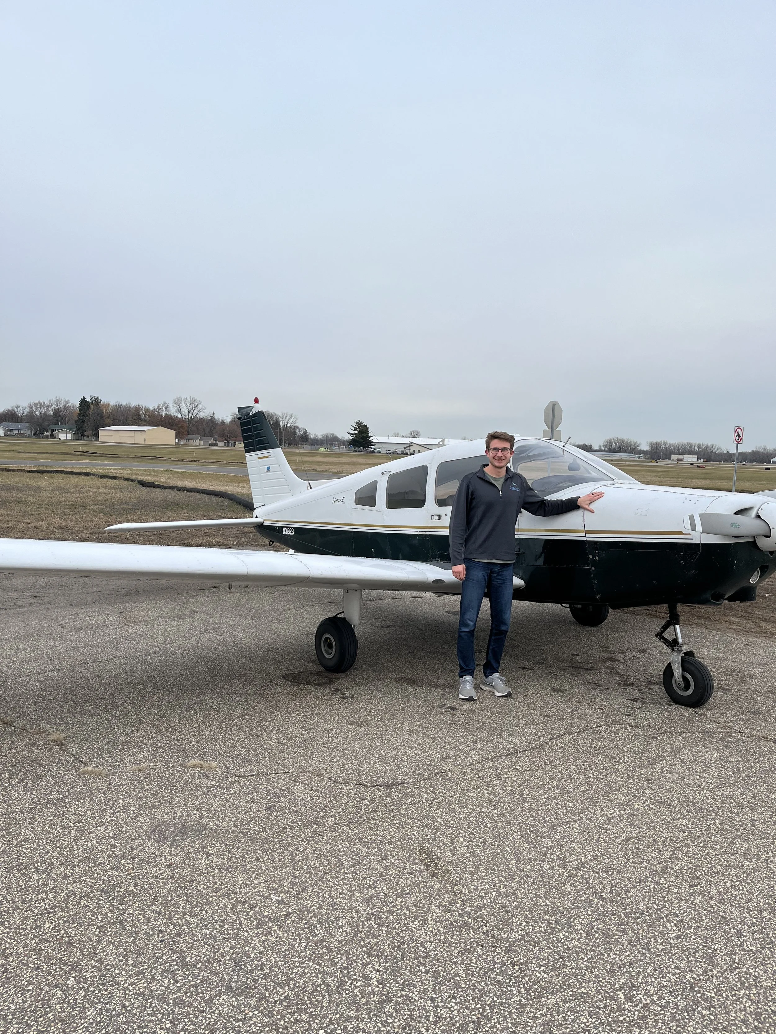 A man standing next to a small airplane on a tarmac, with open fields and overcast sky in the background.