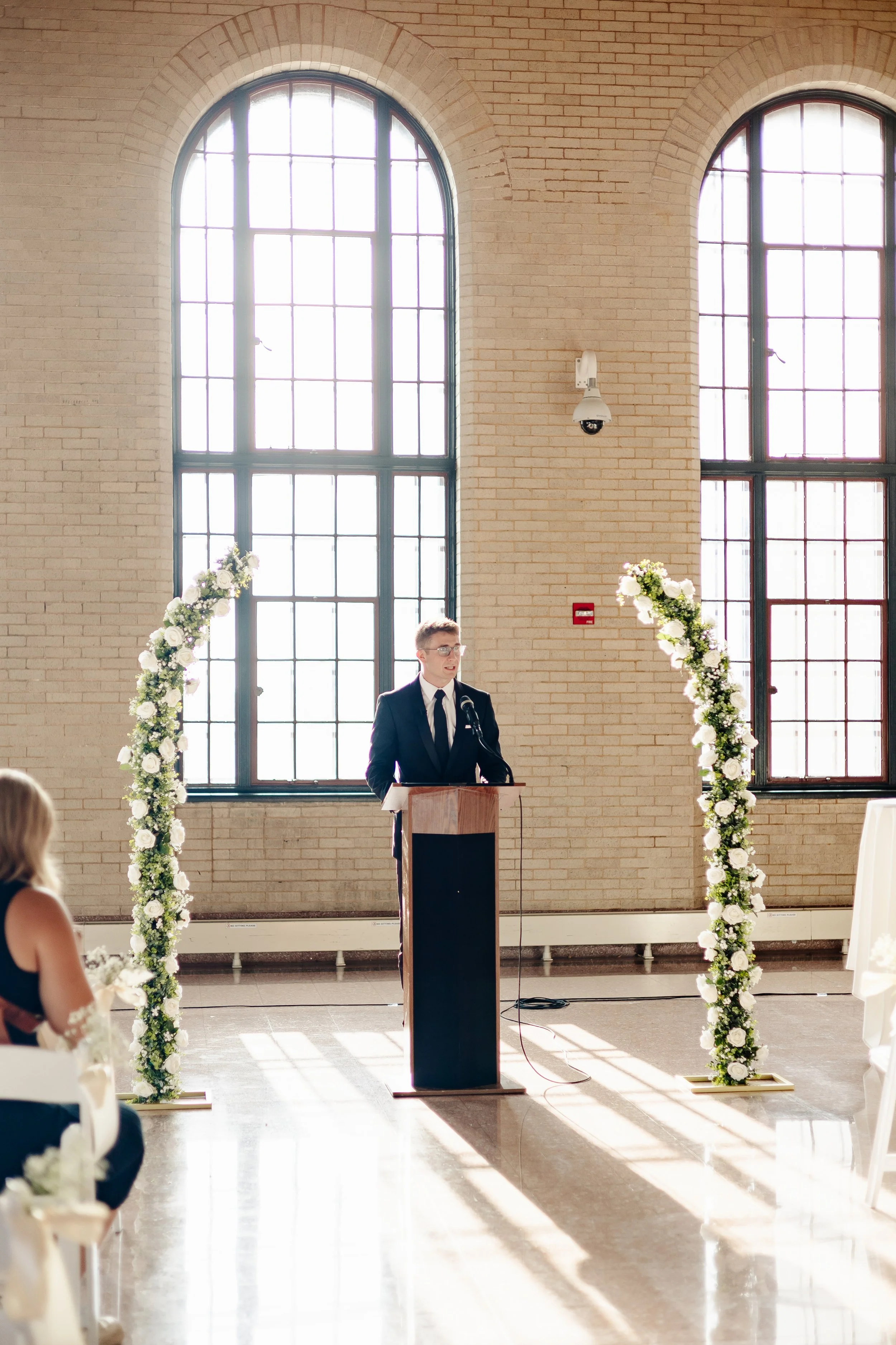 A man in a dark suit and tie giving a speech at a wedding ceremony, standing behind a wooden podium in front of large arched windows and floral arch decorations.