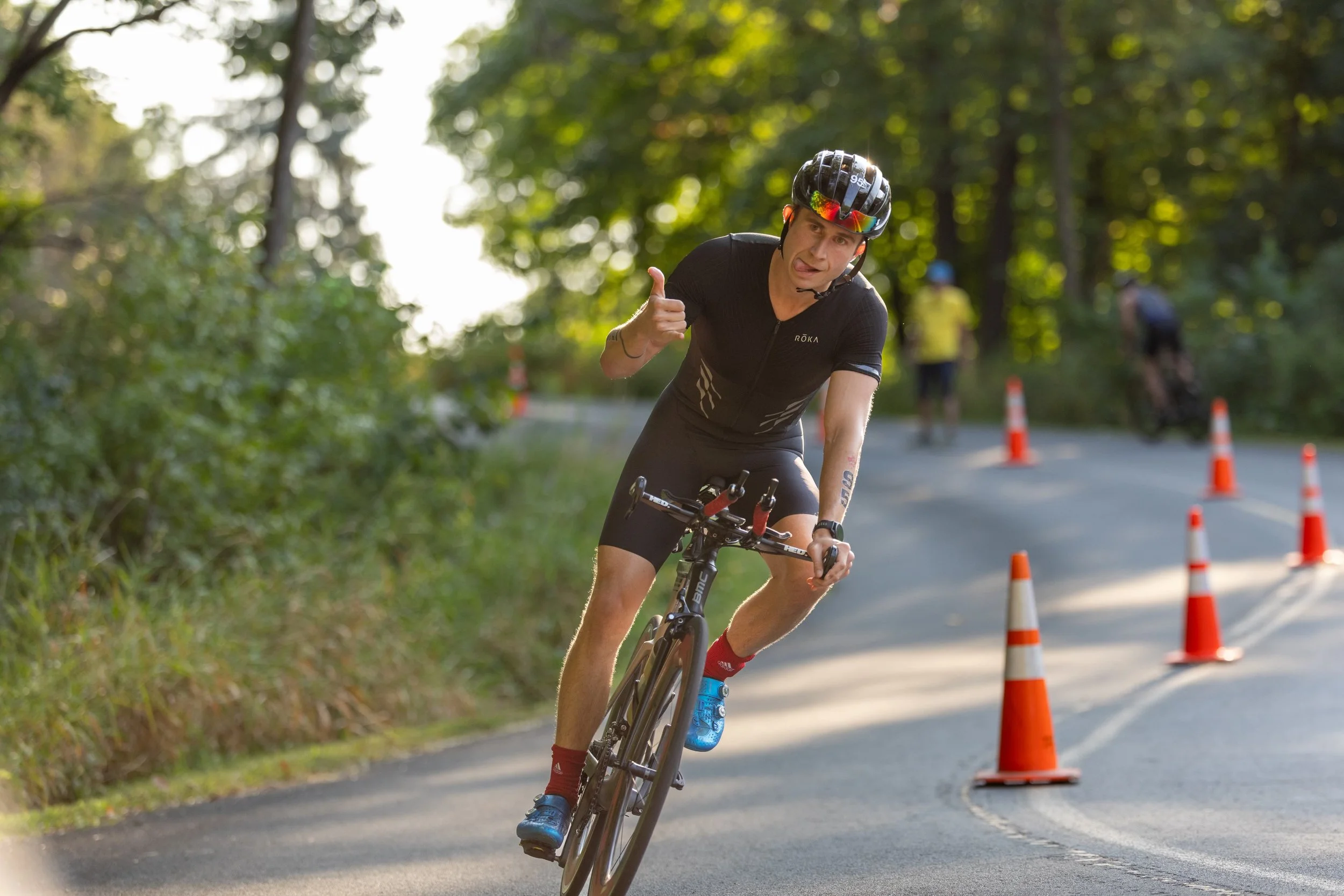 A person riding a bicycle on a mountain road, wearing a helmet and sports gear, giving a thumbs-up gesture. Traffic cones are set up along the curve of the road in a wooded area.