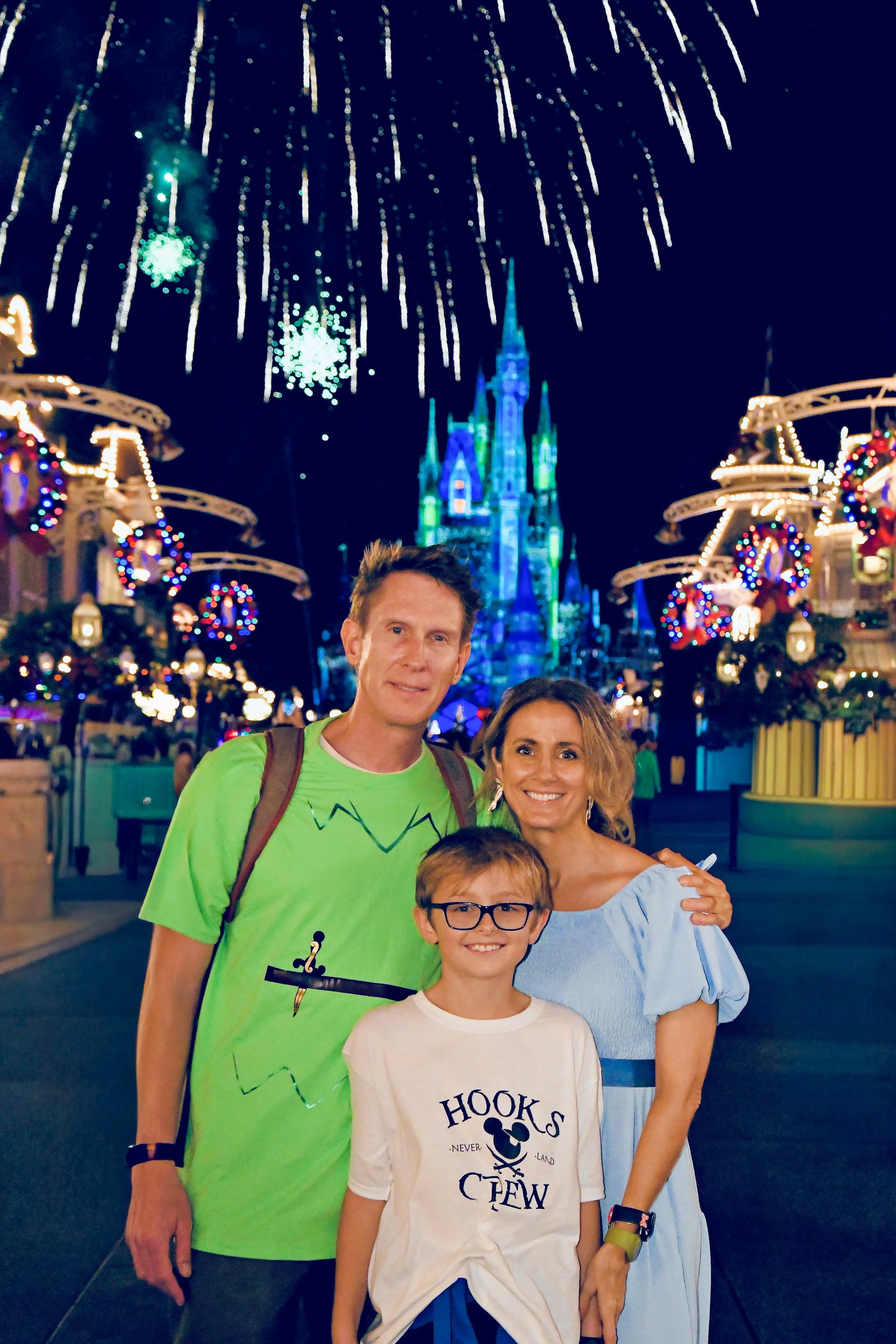 A family of three at a Disney World dressed as Peter Pan, Wendy and Captain Hook Disneybounding with a castle and fireworks in the background, decorated for the holiday season.