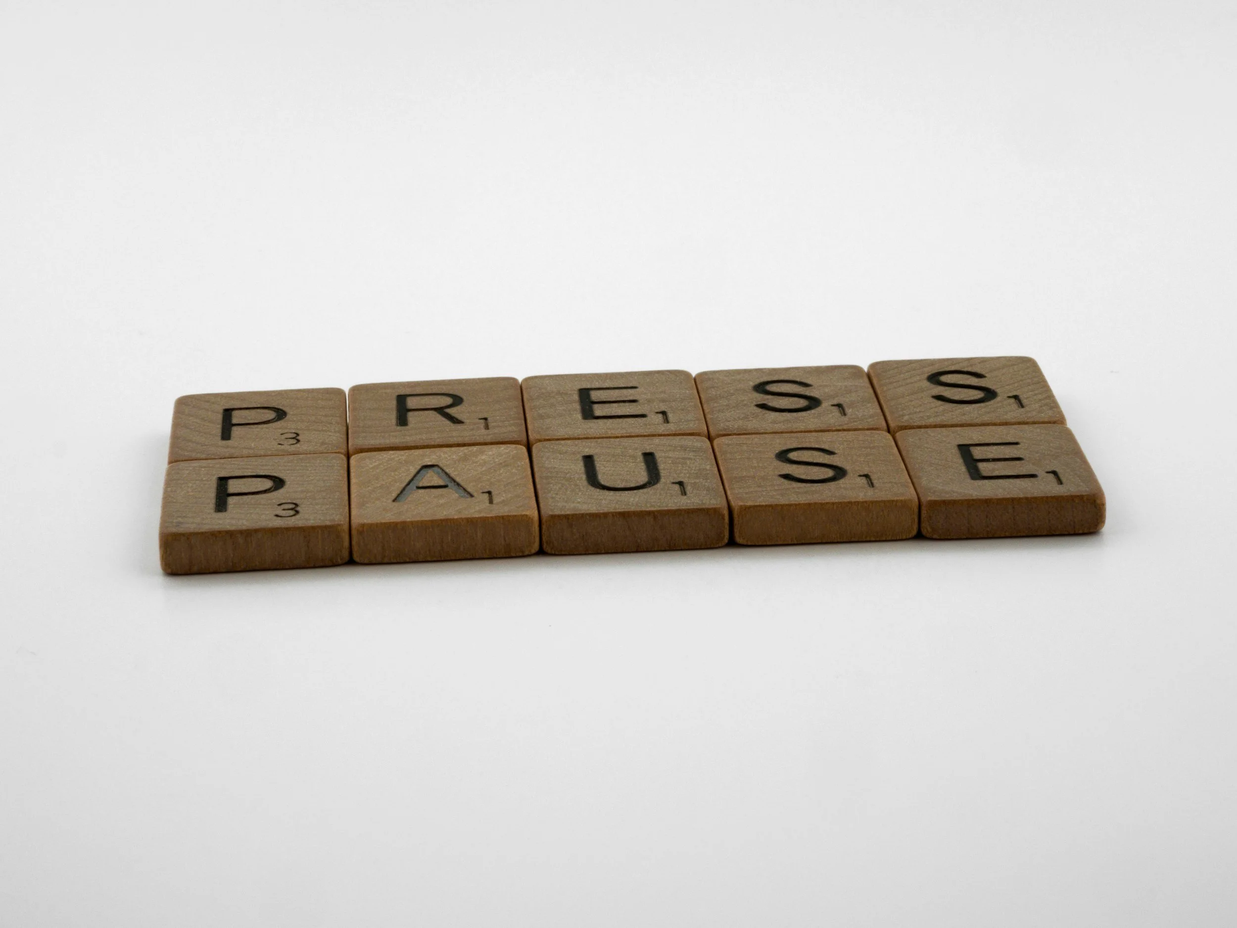 A row of wooden Scrabble tiles spelling the words "PRESS PAUSE" on a plain white background.
