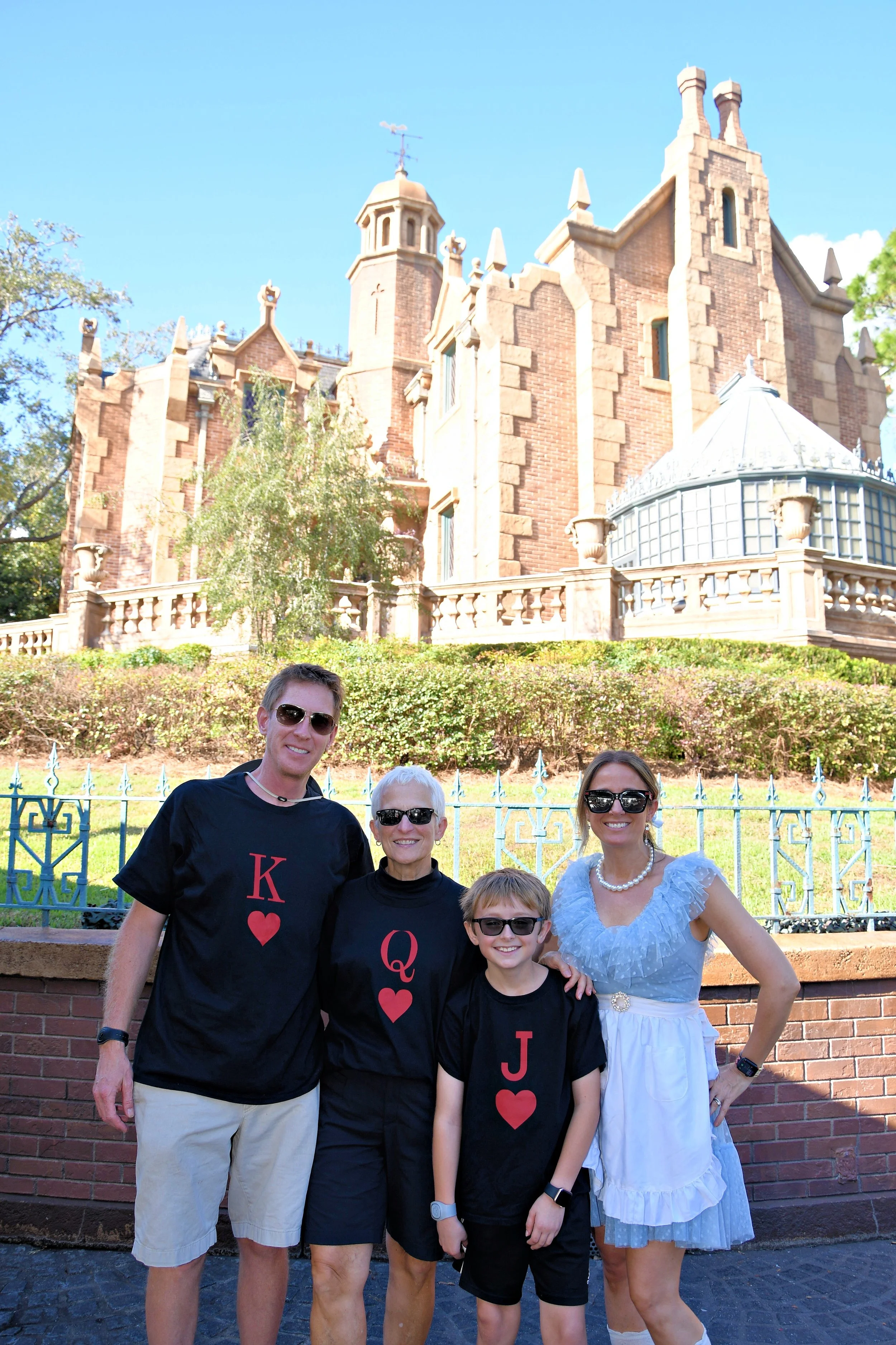 Family of five standing in front of a brick wall with a castle in the background. The family members are wearing matching black shirts with red hearts and letters, and smiling at the camera.  They are dressed as Alice in Wonderland Disneybound