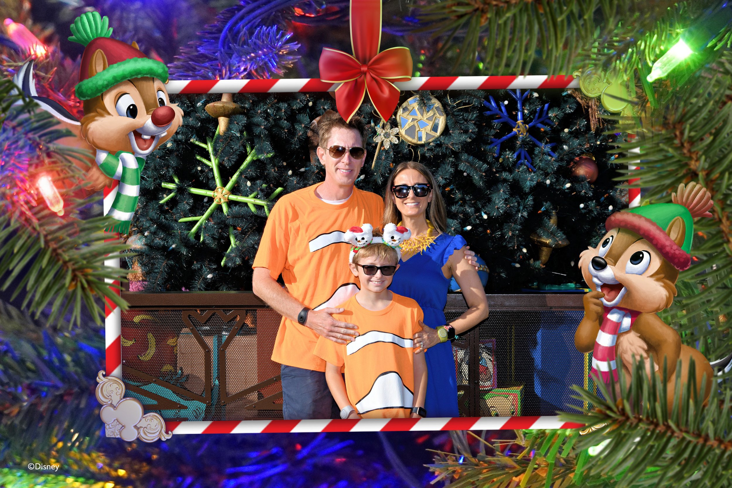 A family posing together at a Christmas celebration, standing in front of a decorated Christmas tree with ornaments. The father, mother, and two children are smiling, with the father and son wearing matching orange and white striped shirts. The scene is framed with artificial pine branches and holiday decorations, including characters from Disney's Chip and Dale.
