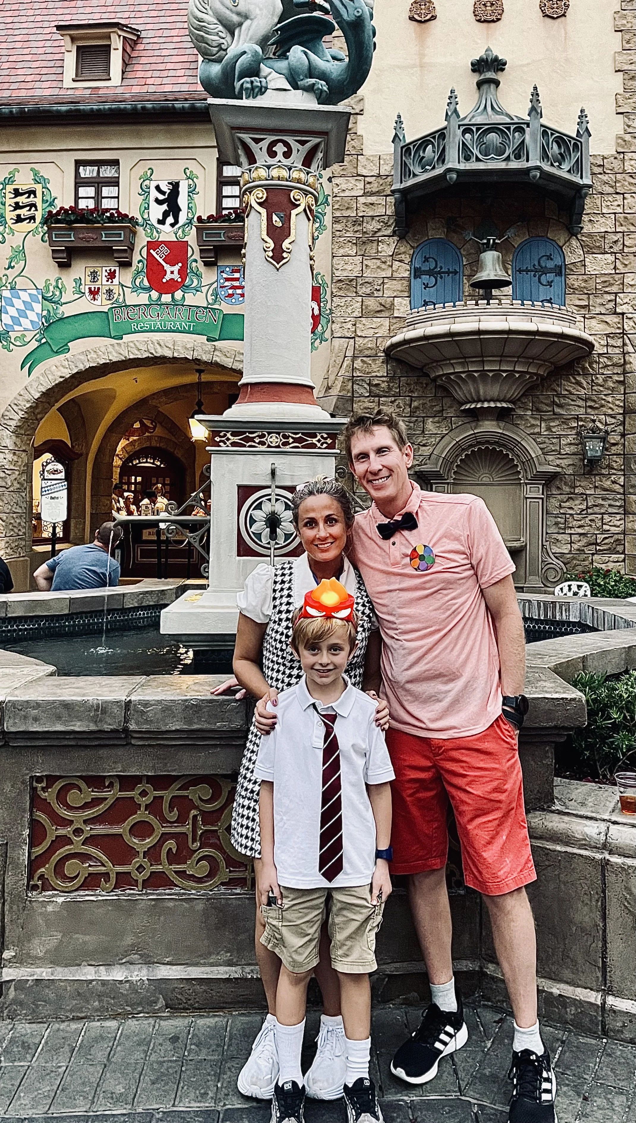 A family of three standing in front of a fountain outside a building with medieval-style architecture, including shields and a balcony with a bell. The family is dressed casually, with the father wearing a pink shirt, the mother in a black and white checkered dress, and the child wearing a white shirt with a striped tie.